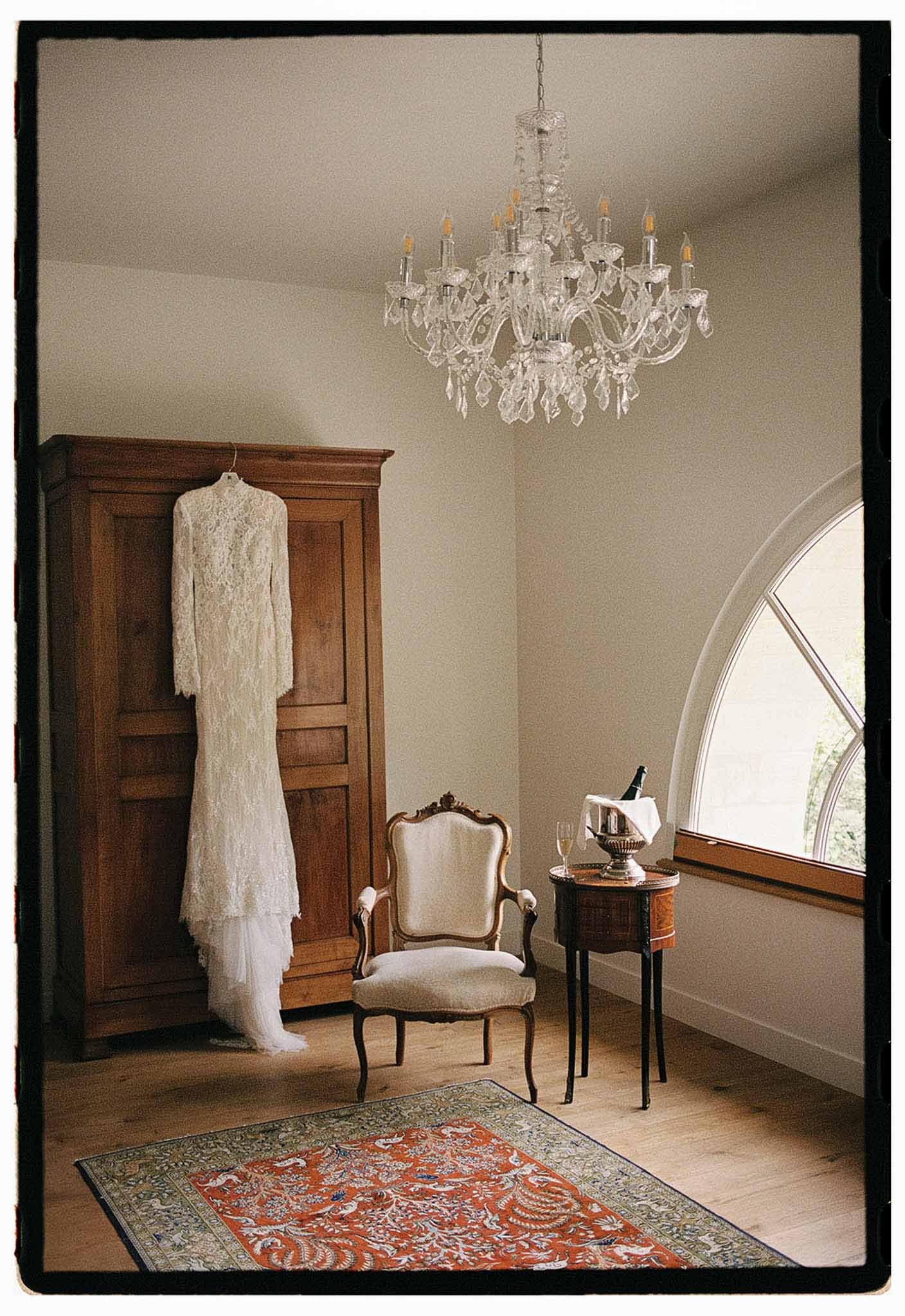 Ivory lace wedding dress hanging on an antique armoire in a chateau room with chandelier and champagne on side table