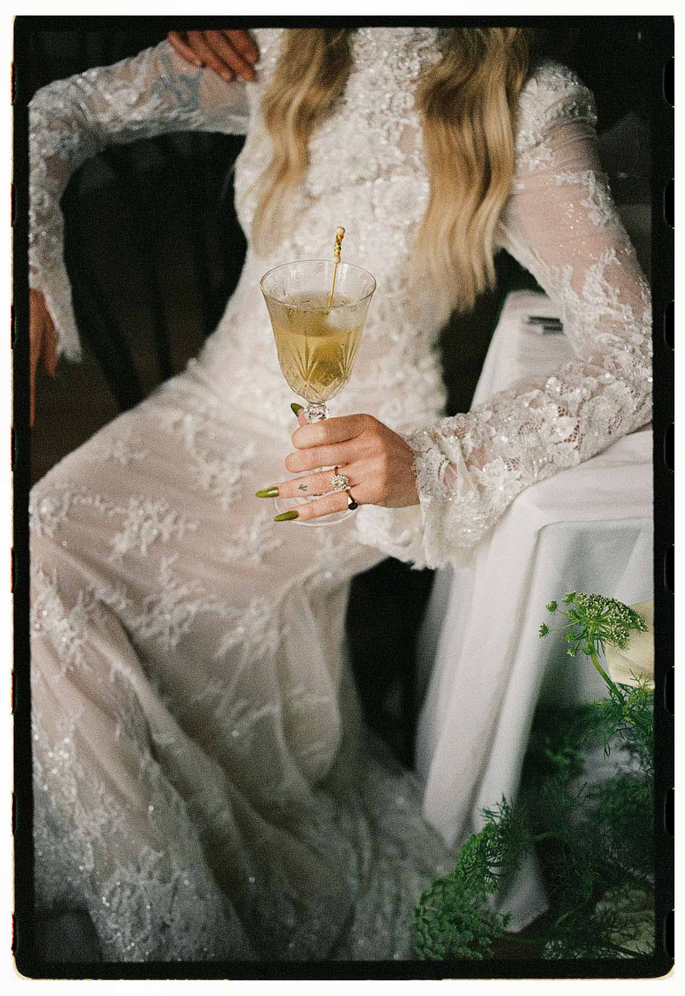Close-up bride holding pale green cocktail in crystal coupe, lace sleeve detail with diamond ring and tattoo