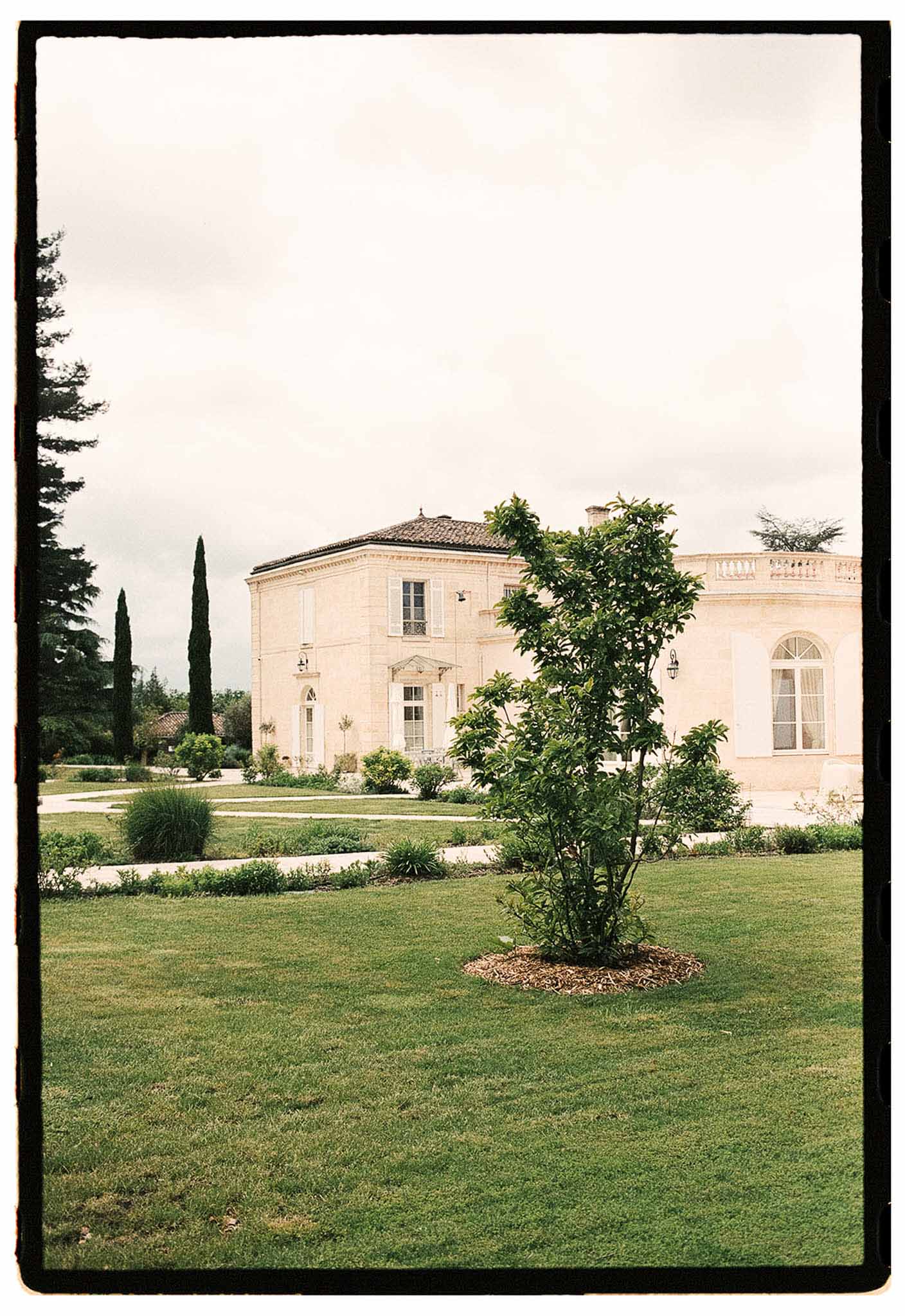 Film-grain shot of cream limestone chateau with dark tile roof, balustrade wing, and manicured lawn