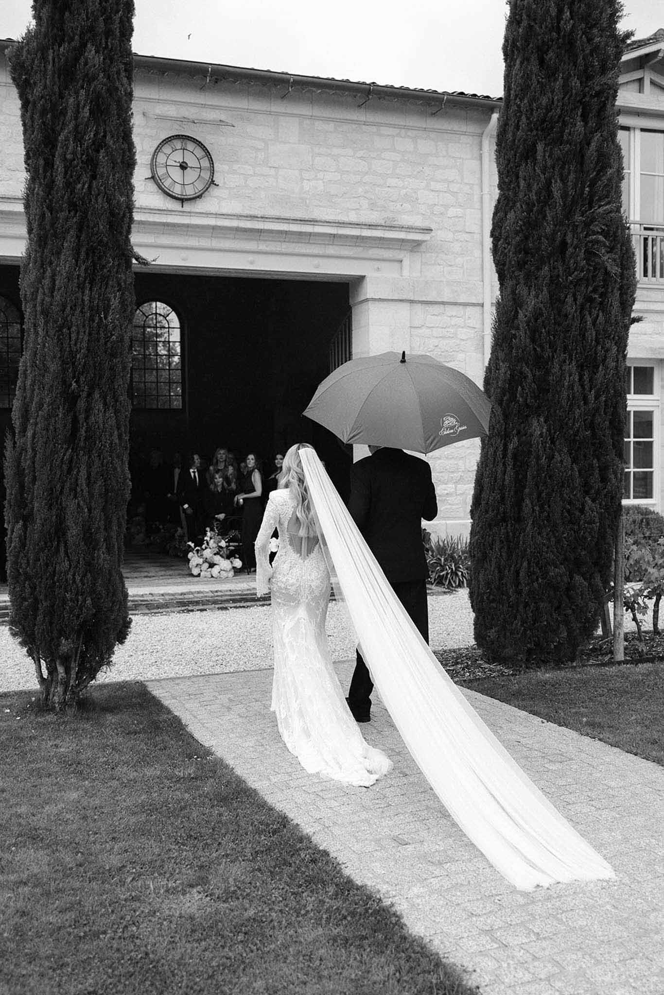 Black and white of bride with cathedral veil escorted under umbrella along cypress-lined path toward stone venue