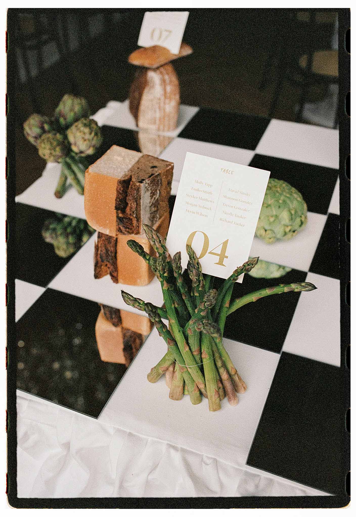 Escort card display using asparagus artichokes and cheese as holders on checkered surface