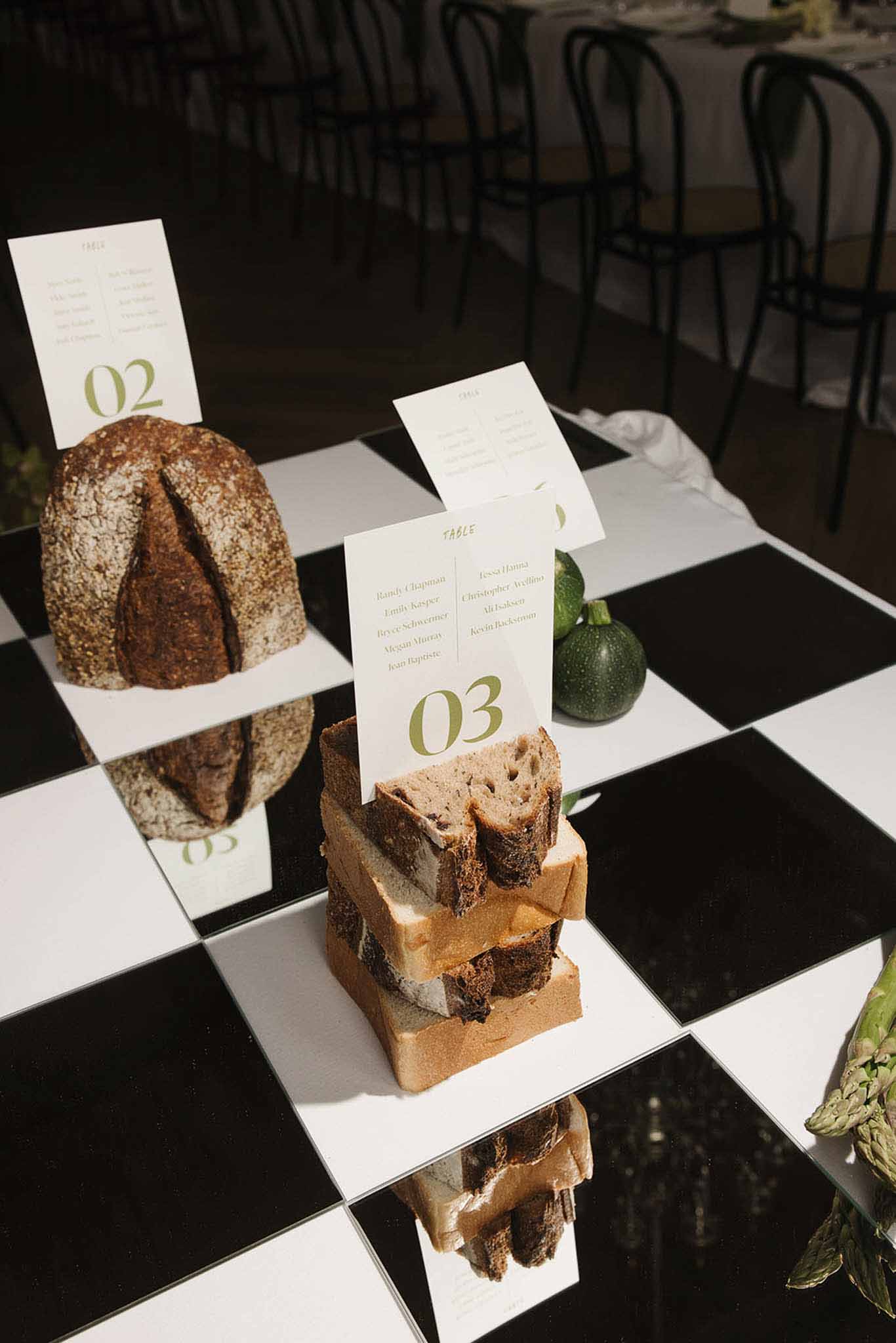 Artisan bread loaves holding olive-green table number cards with courgettes on checkered mirror surface