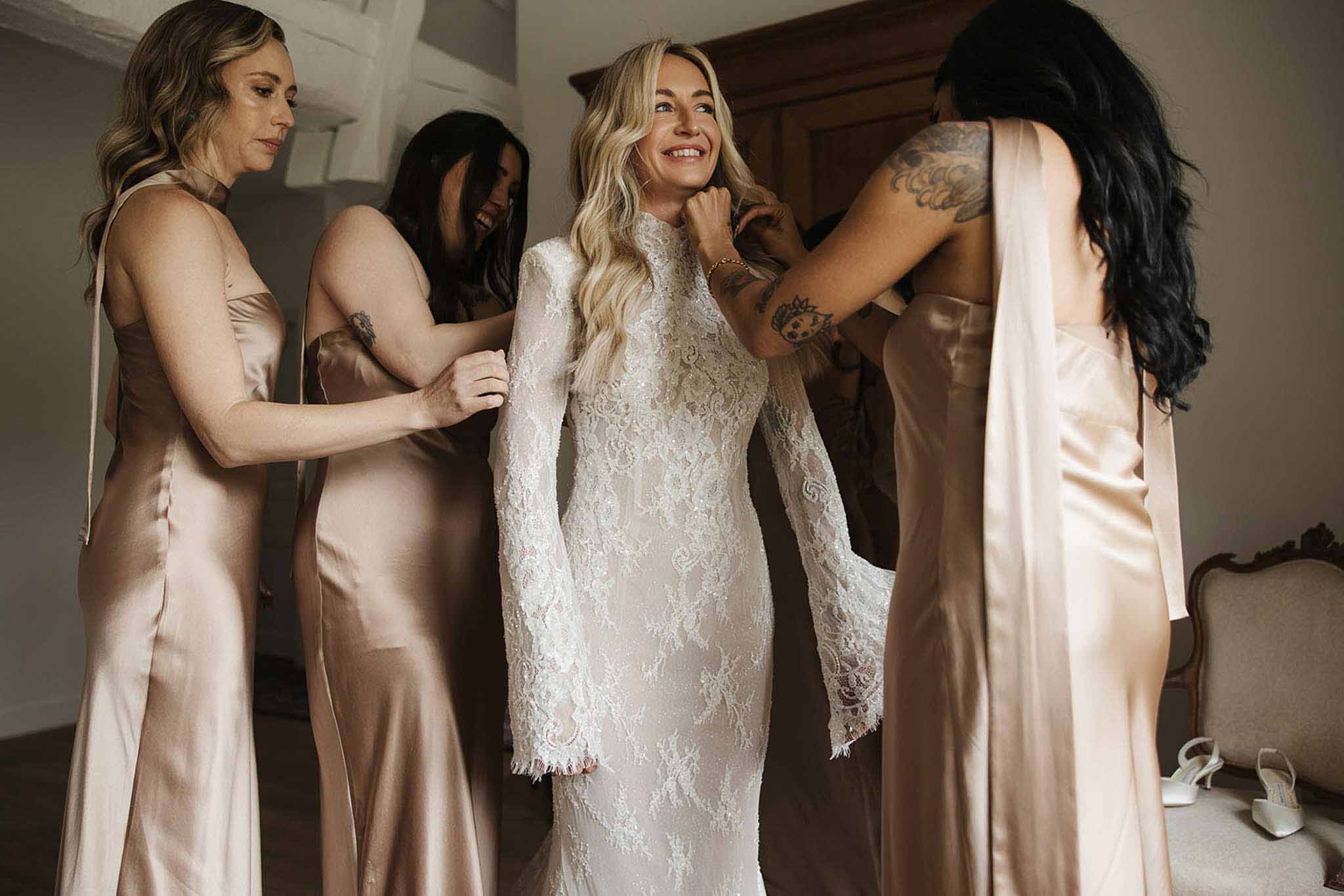 Three bridesmaids in champagne satin dresses helping bride into high-neck lace gown in chateau room