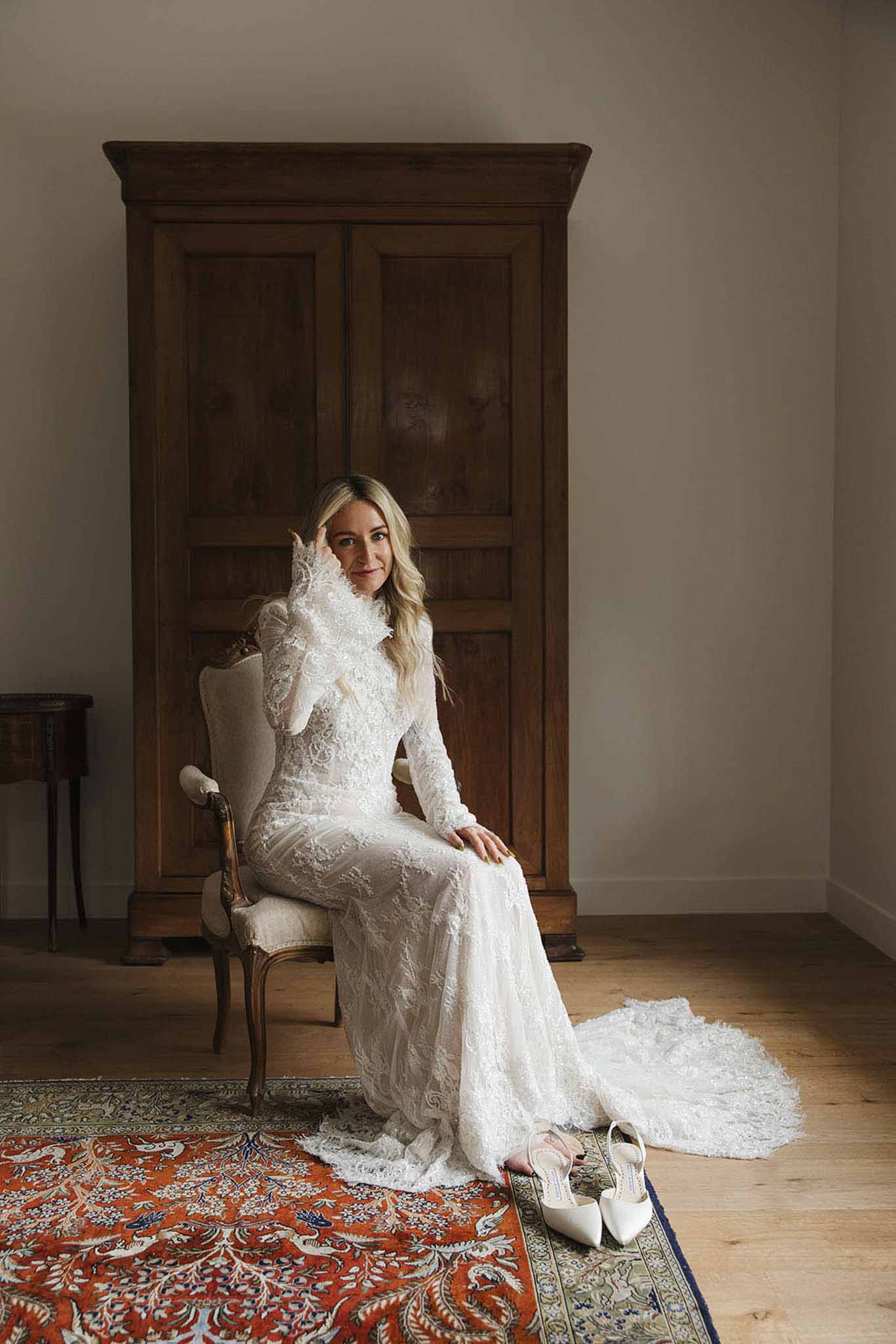 Bride in long-sleeve ruffled lace gown seated on French armchair in chateau room