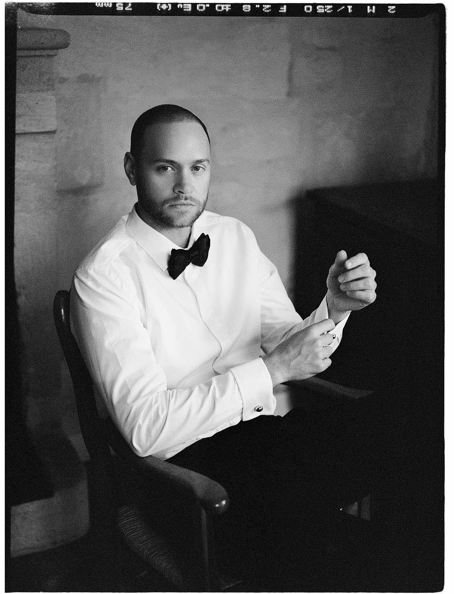 Black-and-white portrait of groom fastening cufflinks while seated in chair wearing white shirt and bow tie