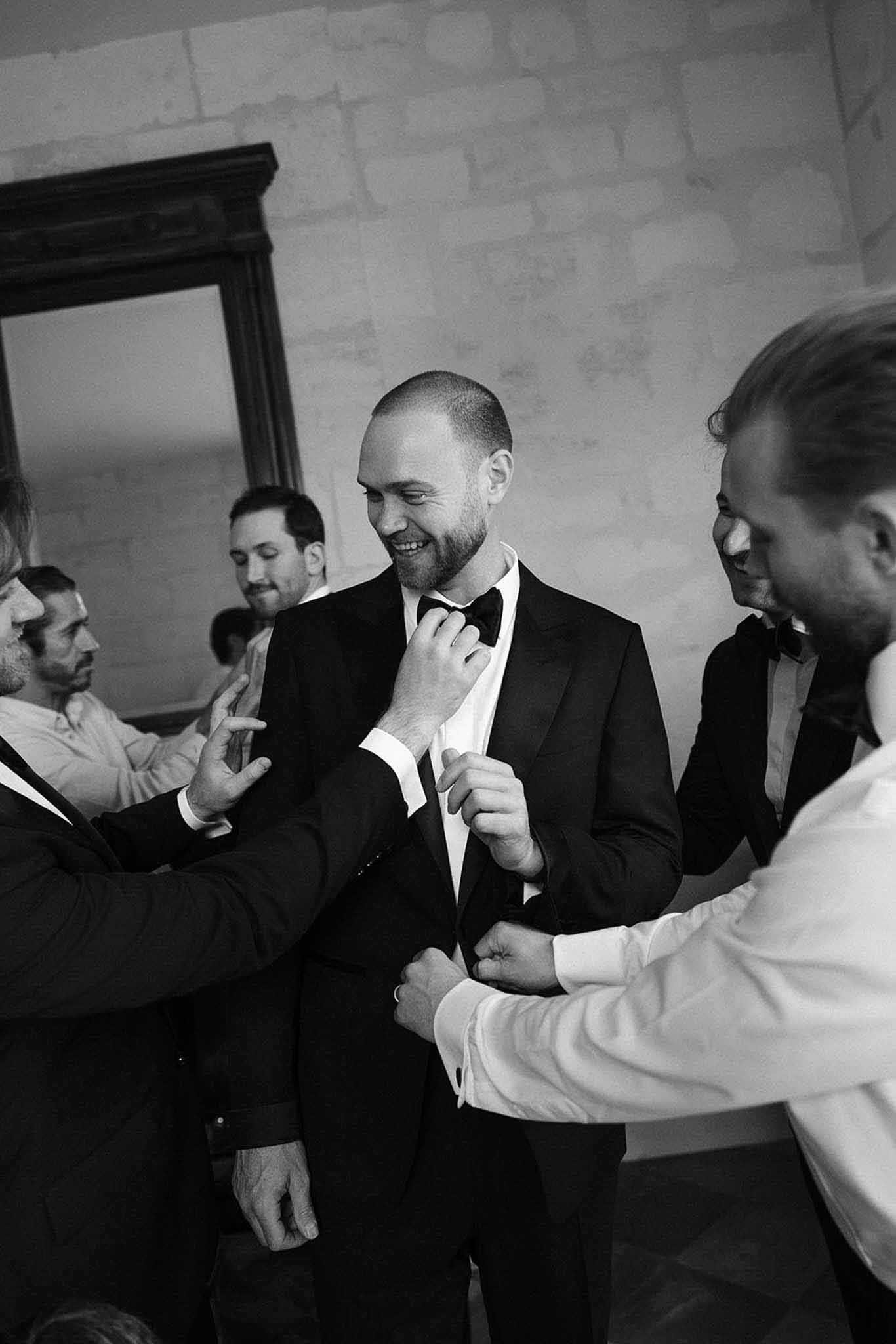 Black and white candid of groomsmen adjusting groom's tuxedo and bow tie in stone-walled room