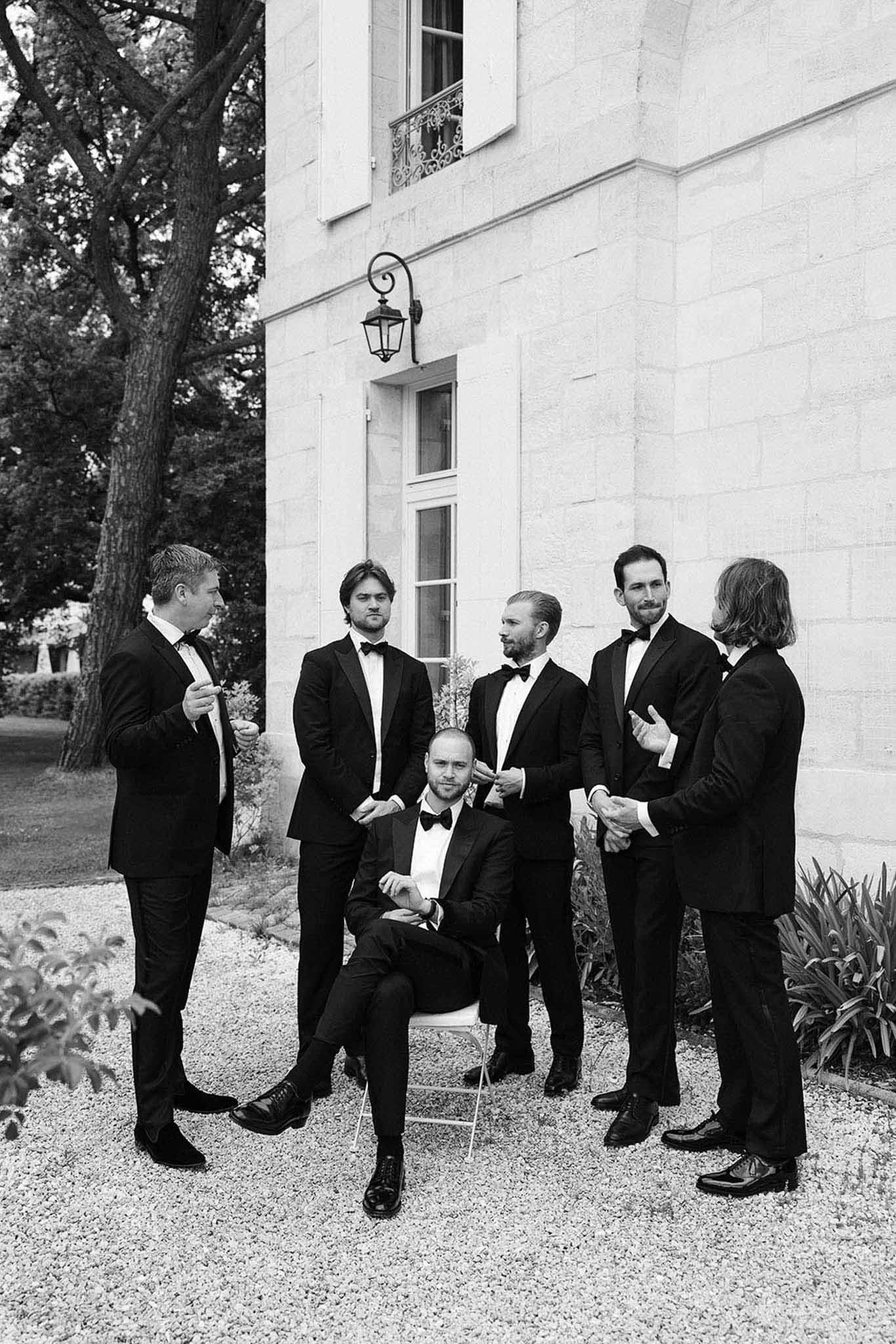 Black and white portrait of groom seated with five groomsmen in matching black tuxedos outside French chateau