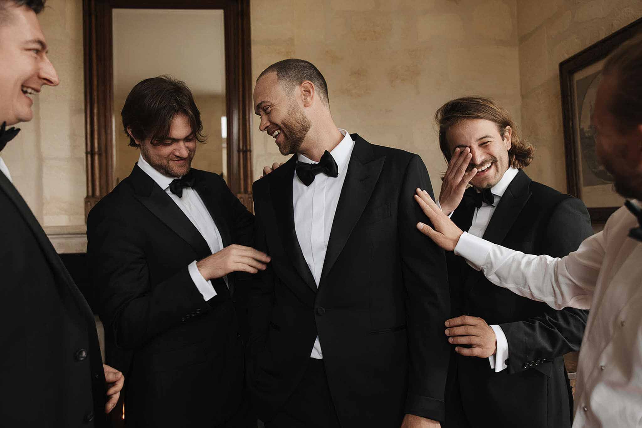 Groom and three groomsmen in black tuxedos laughing together in chateau interior room