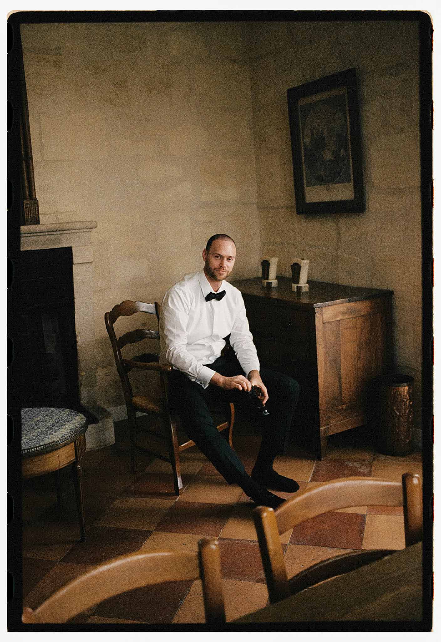 Groom in white shirt and bow tie adjusting shoes in stone-walled chateau room with antique furniture
