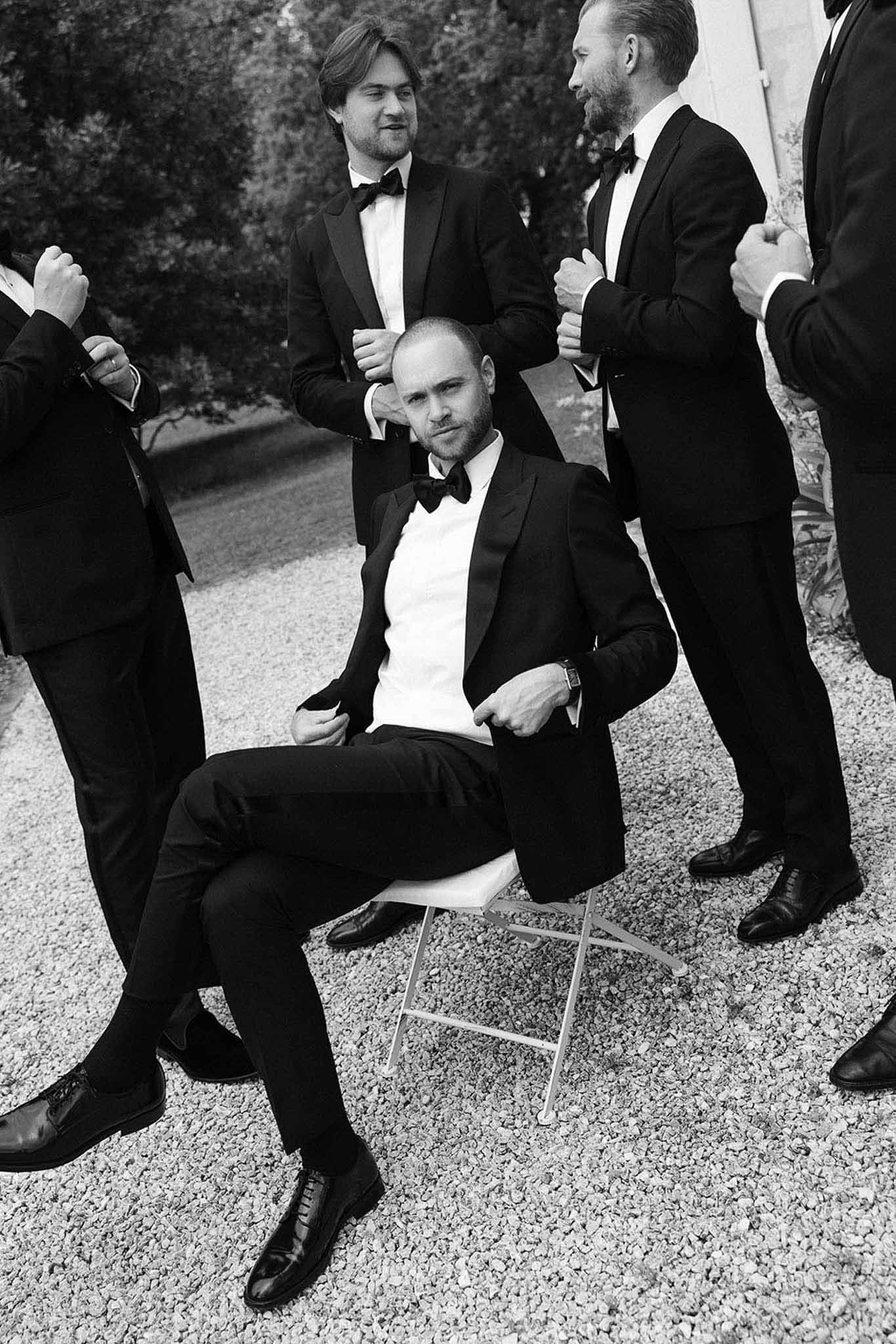 Black and white groom seated with four groomsmen standing in matching tuxedos in chateau courtyard