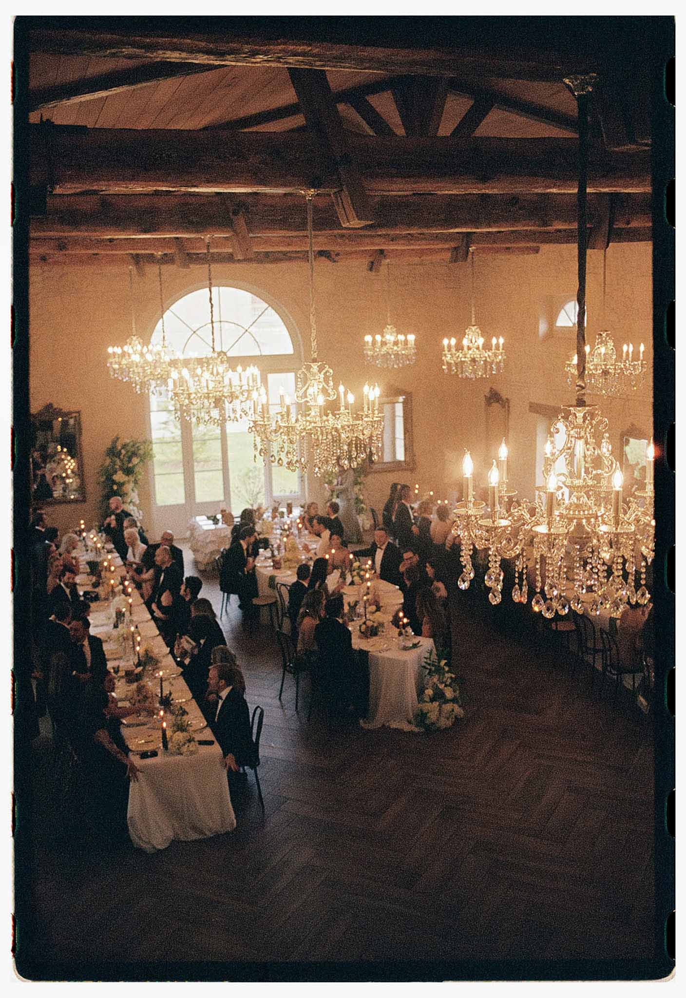 Elevated view of formal reception dinner in chateau ballroom with chandeliers, candles, and 50 seated guests