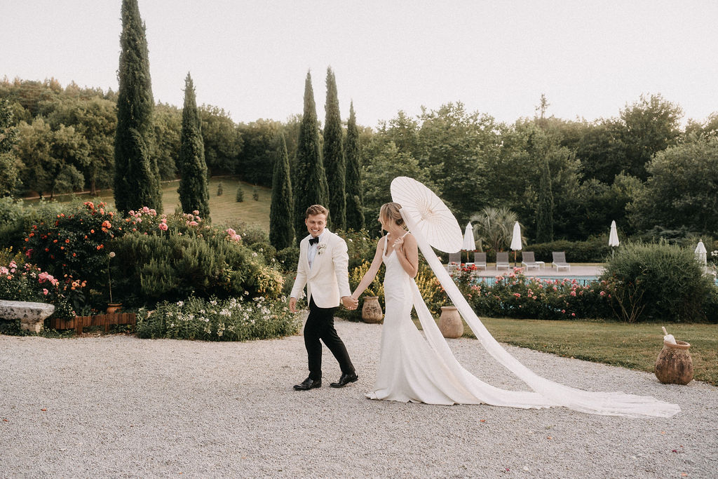 Groom leading bride with parasol and cathedral train past cypress trees and rose garden at estate