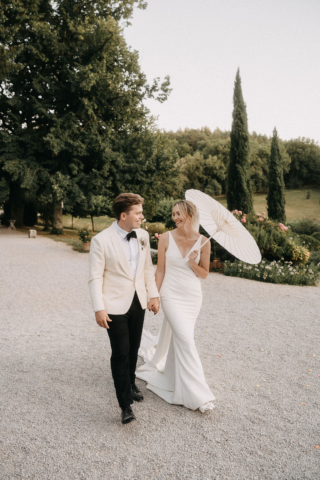 Bride with paper parasol and groom in cream dinner jacket walk hand-in-hand on estate gravel path