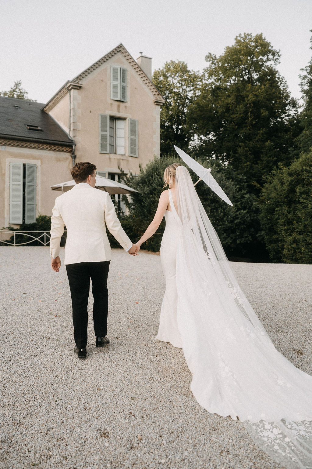 Bride with parasol and applique veil and groom in ivory blazer walk toward country house from behind