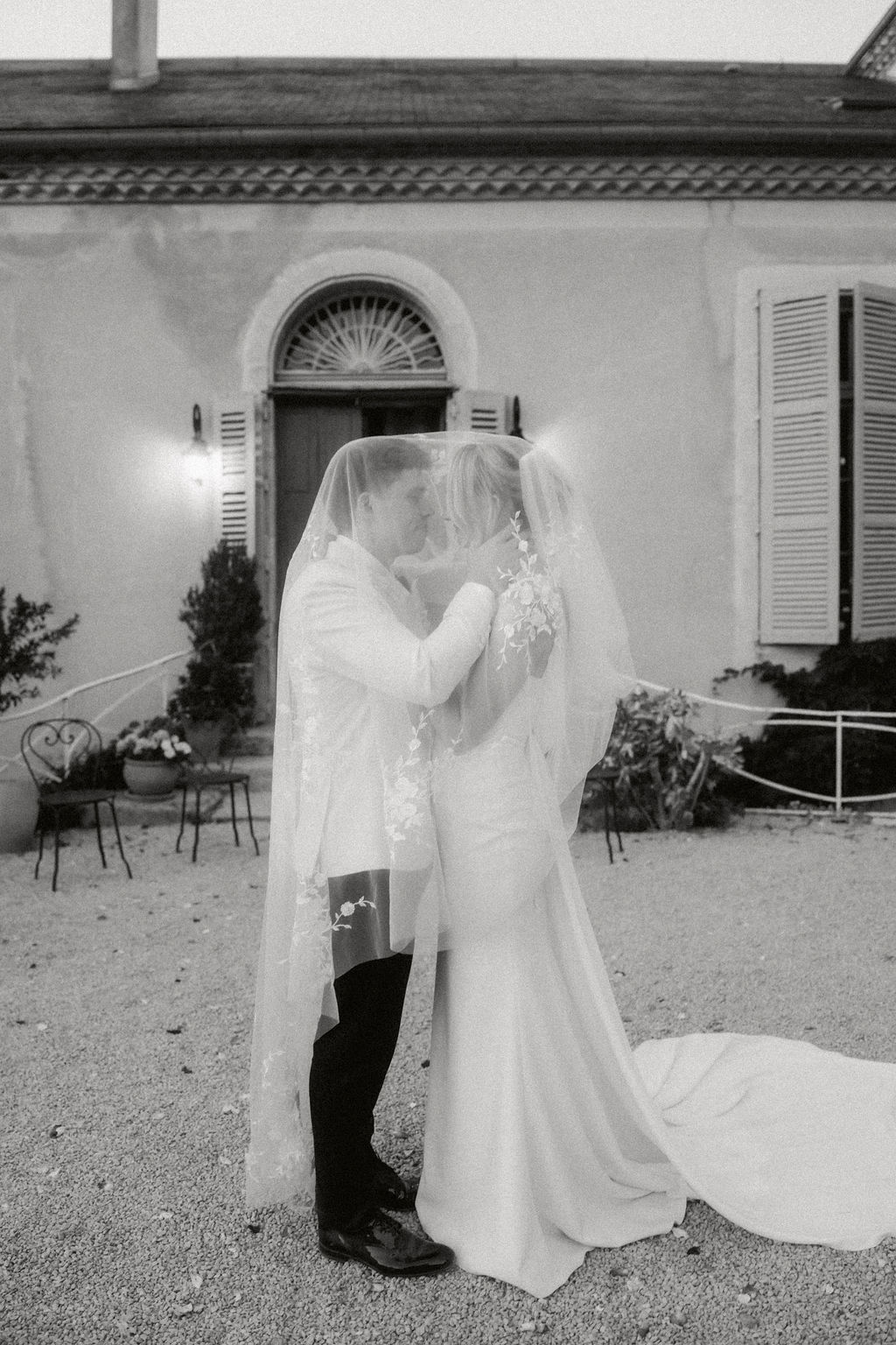 Black and white couple under cathedral veil with floral trim on lit manor house gravel courtyard at dusk