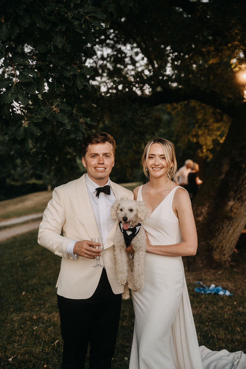 Bride and groom in garden at golden hour holding small white dog dressed in tuxedo bandana