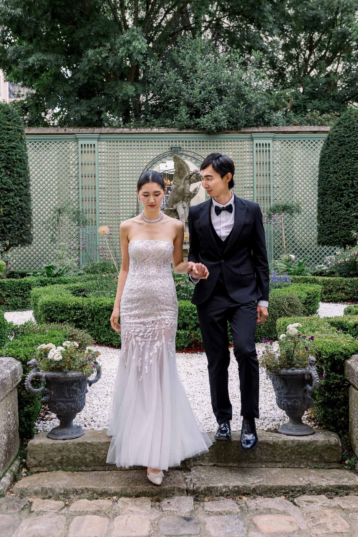 Bride in beaded mermaid gown and groom descending stone steps in formal boxwood garden