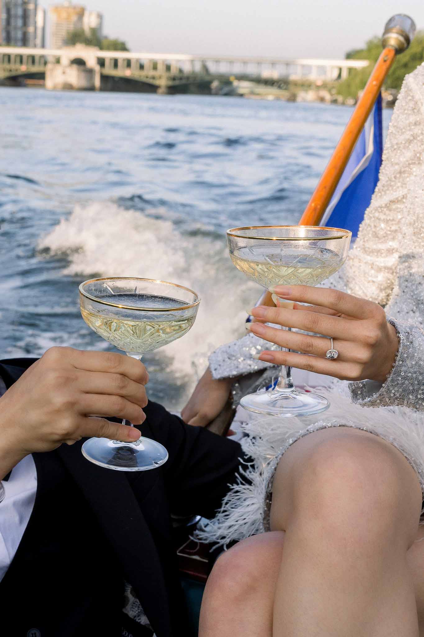 Couple holding gold-rimmed champagne coupes on Seine River boat with Paris bridge backdrop