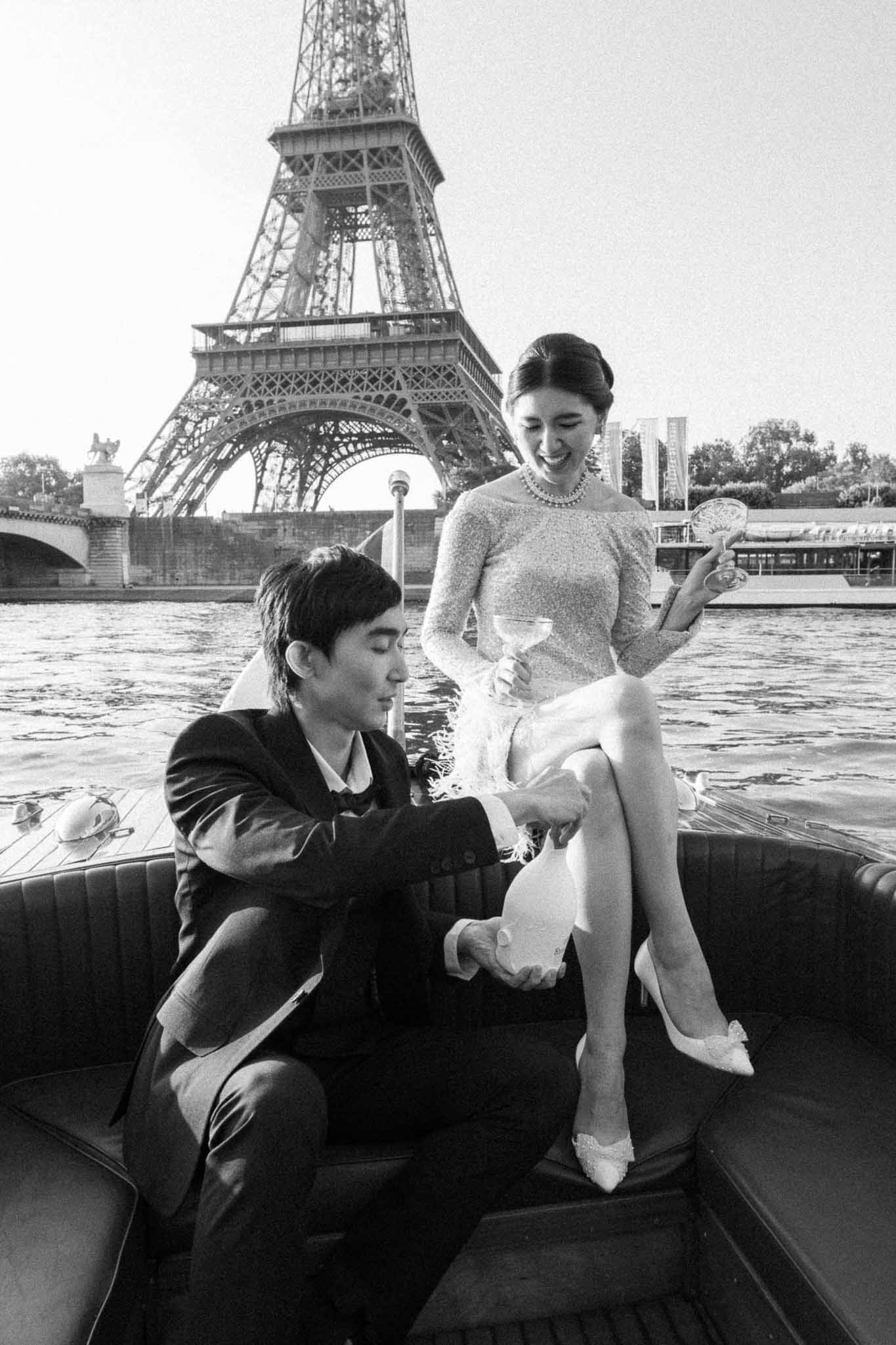 Black-and-white portrait of couple on Seine River boat with Eiffel Tower, groom adjusting bride's shoe