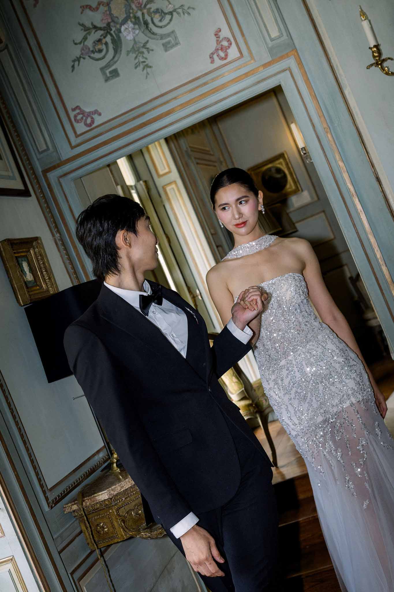 Couple holding hands in ornate chateau doorway, bride in beaded silver halter gown, groom in black tuxedo