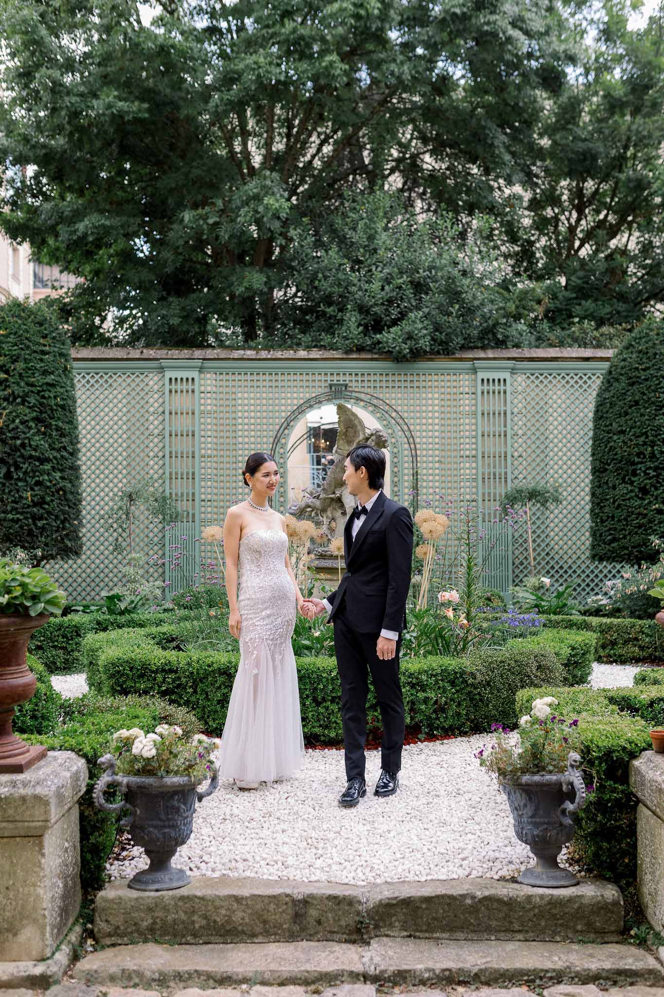 Couple holds hands on gravel path in formal garden with boxwood parterres and sage lattice trellis behind