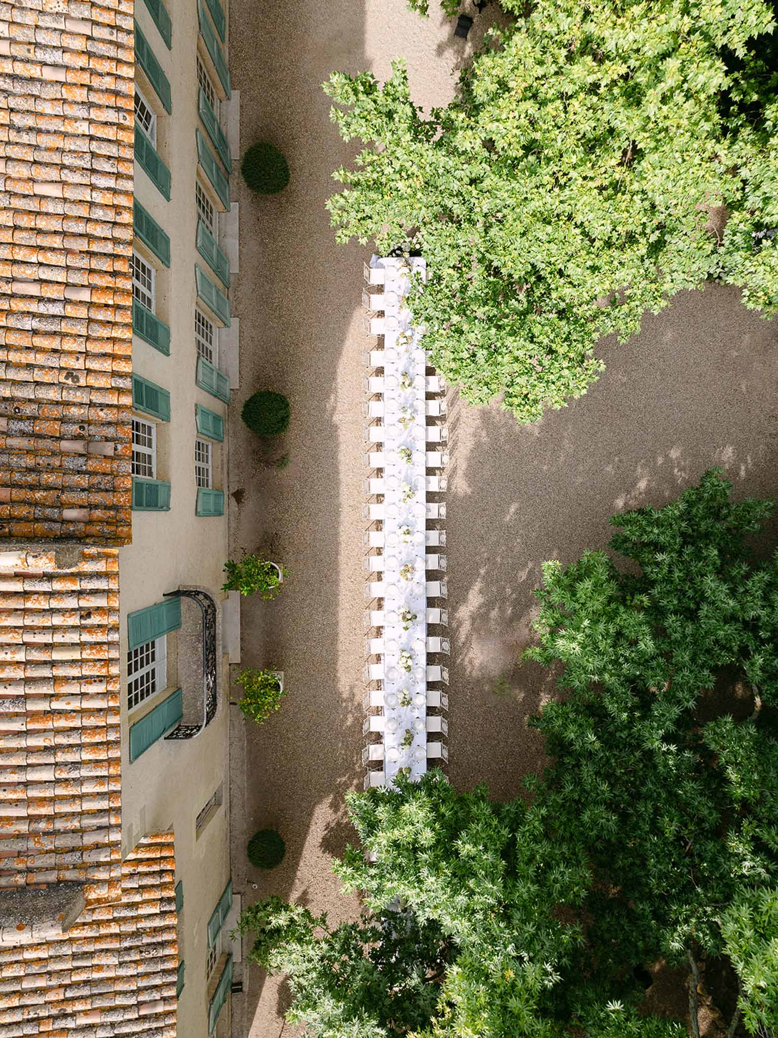 Aerial view of a long banquet table set for 50 guests in a chateau courtyard with topiary and teal shutters