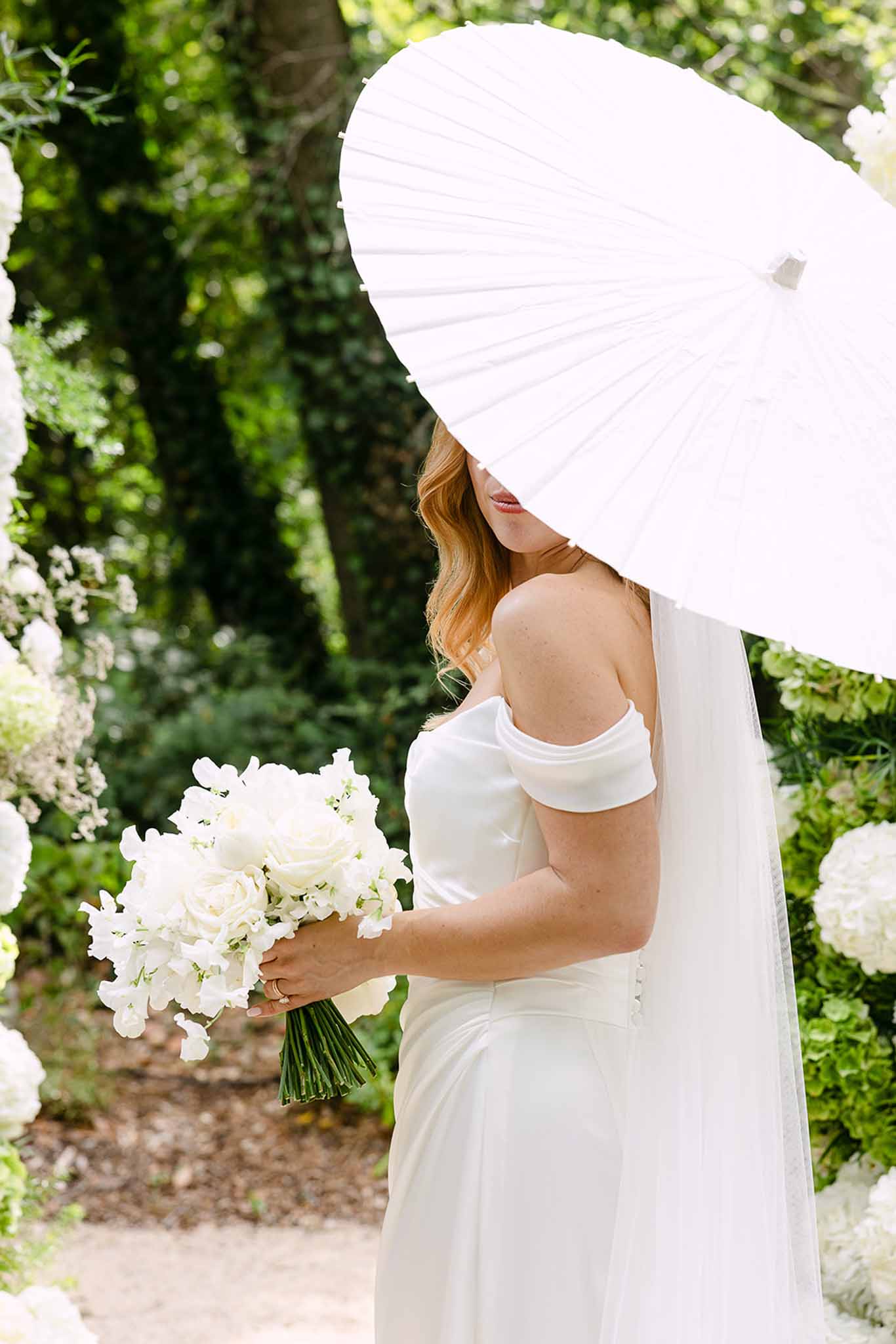 Bride with off-the-shoulder satin gown holding white rose bouquet and paper parasol among hydrangea arrangements