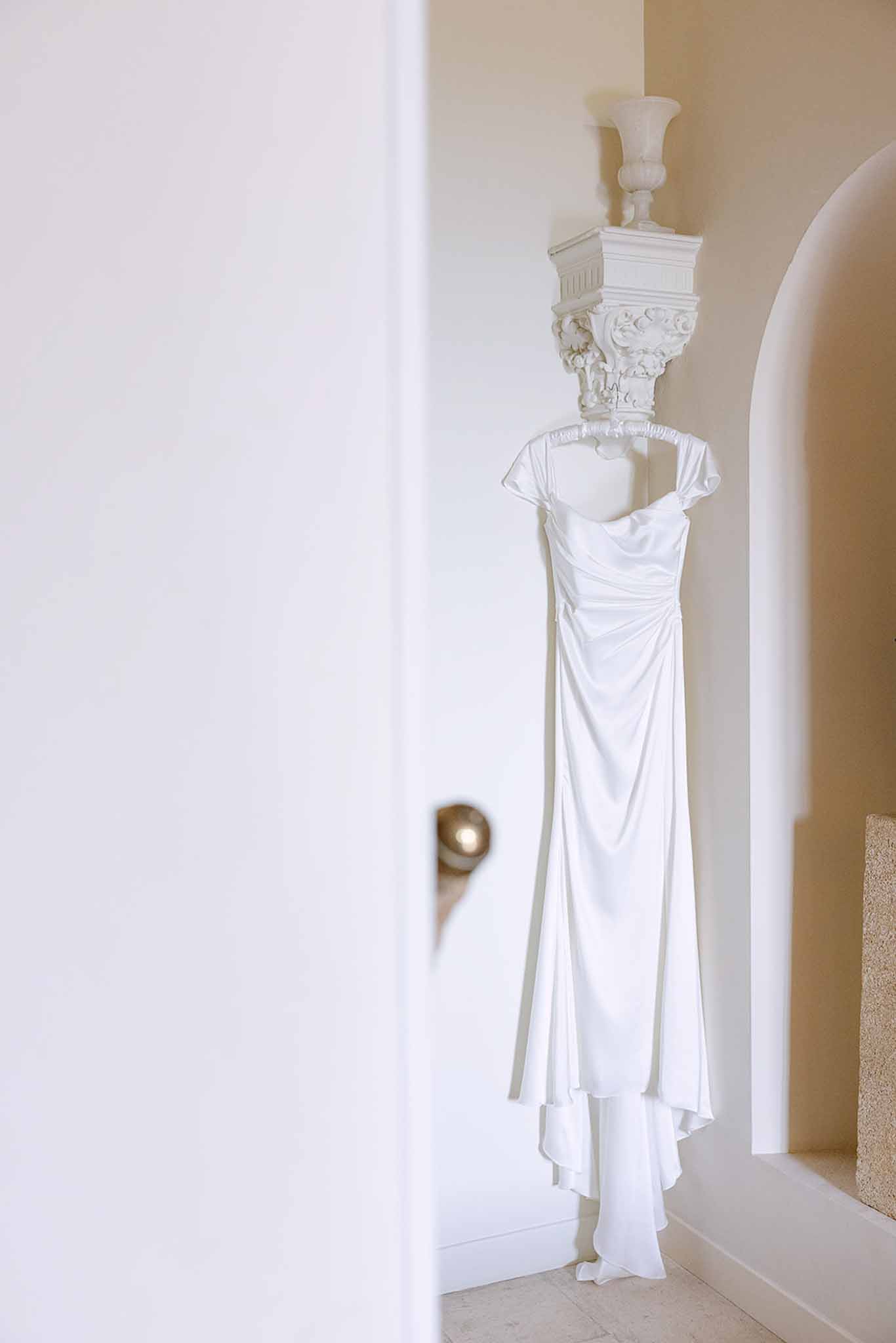 Ivory draped cowl wedding dress hanging from white wall sconce bracket in arched alcove