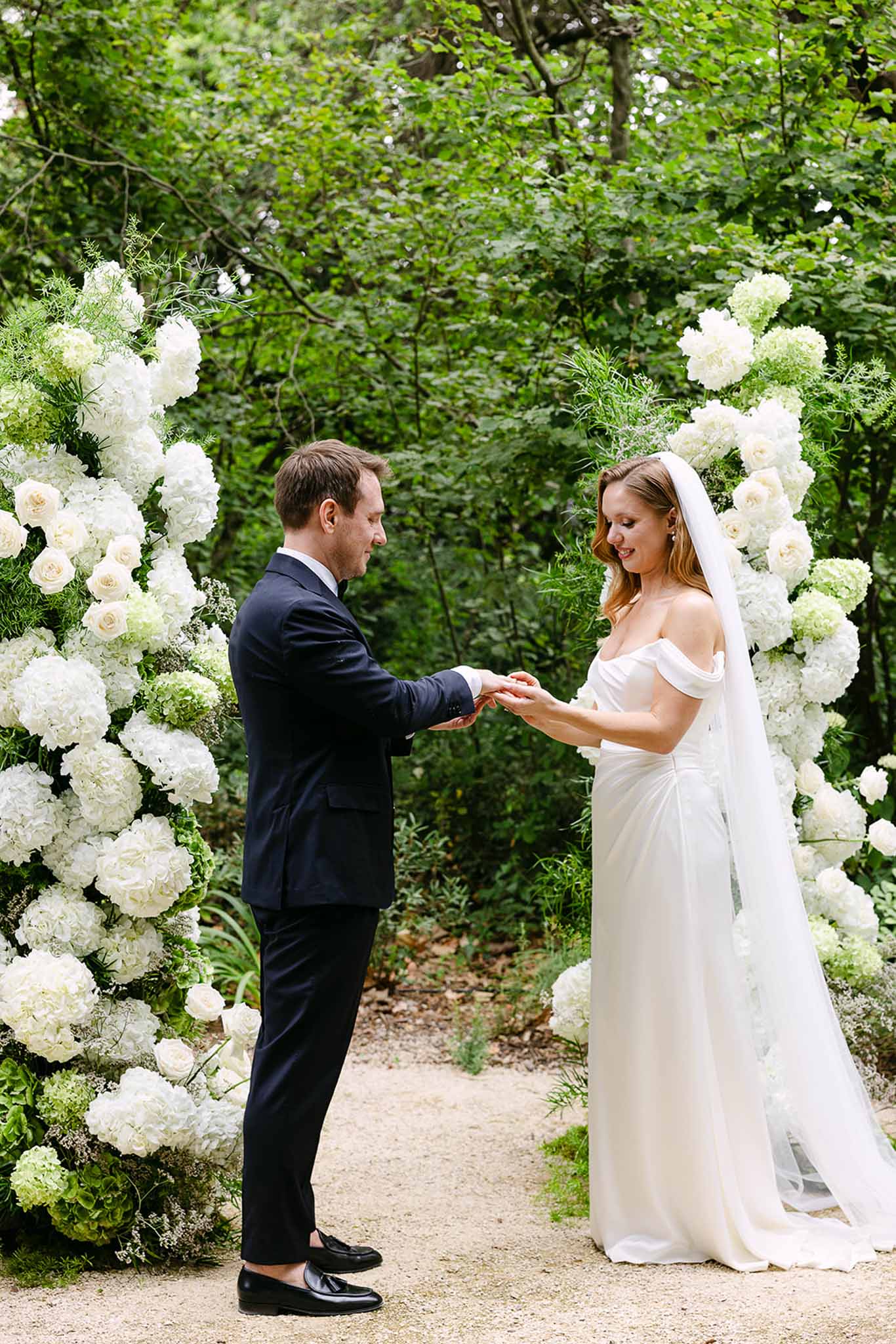 Ring exchange under white hydrangea and rose ceremony arch on gravel path