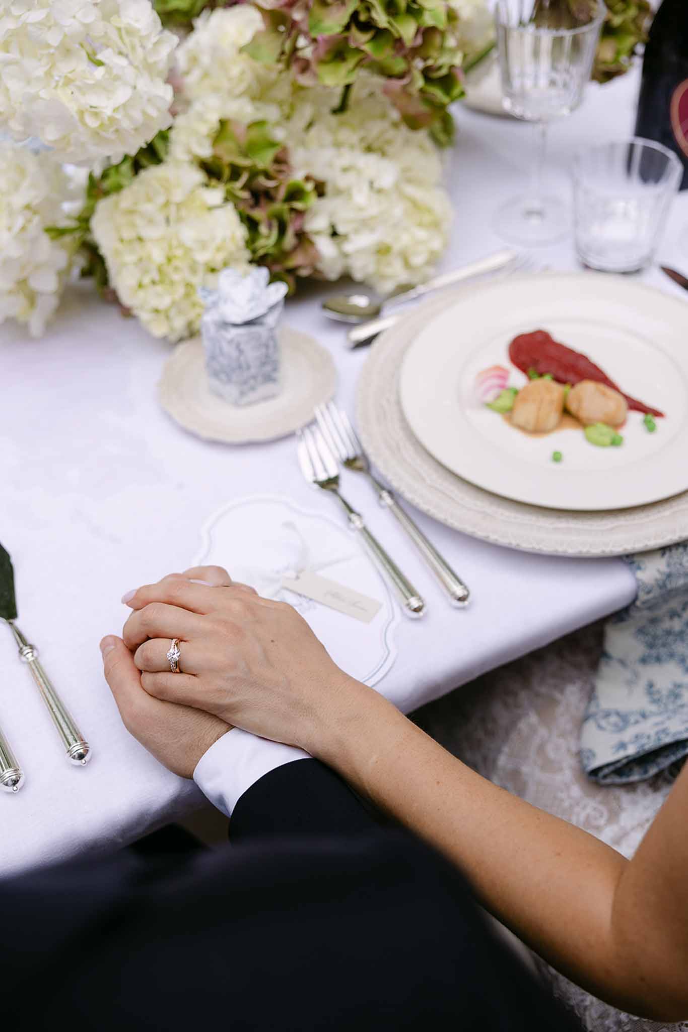 Clasped hands over seared scallop starter with woven charger, toile napkin, and white hydrangea centerpiece