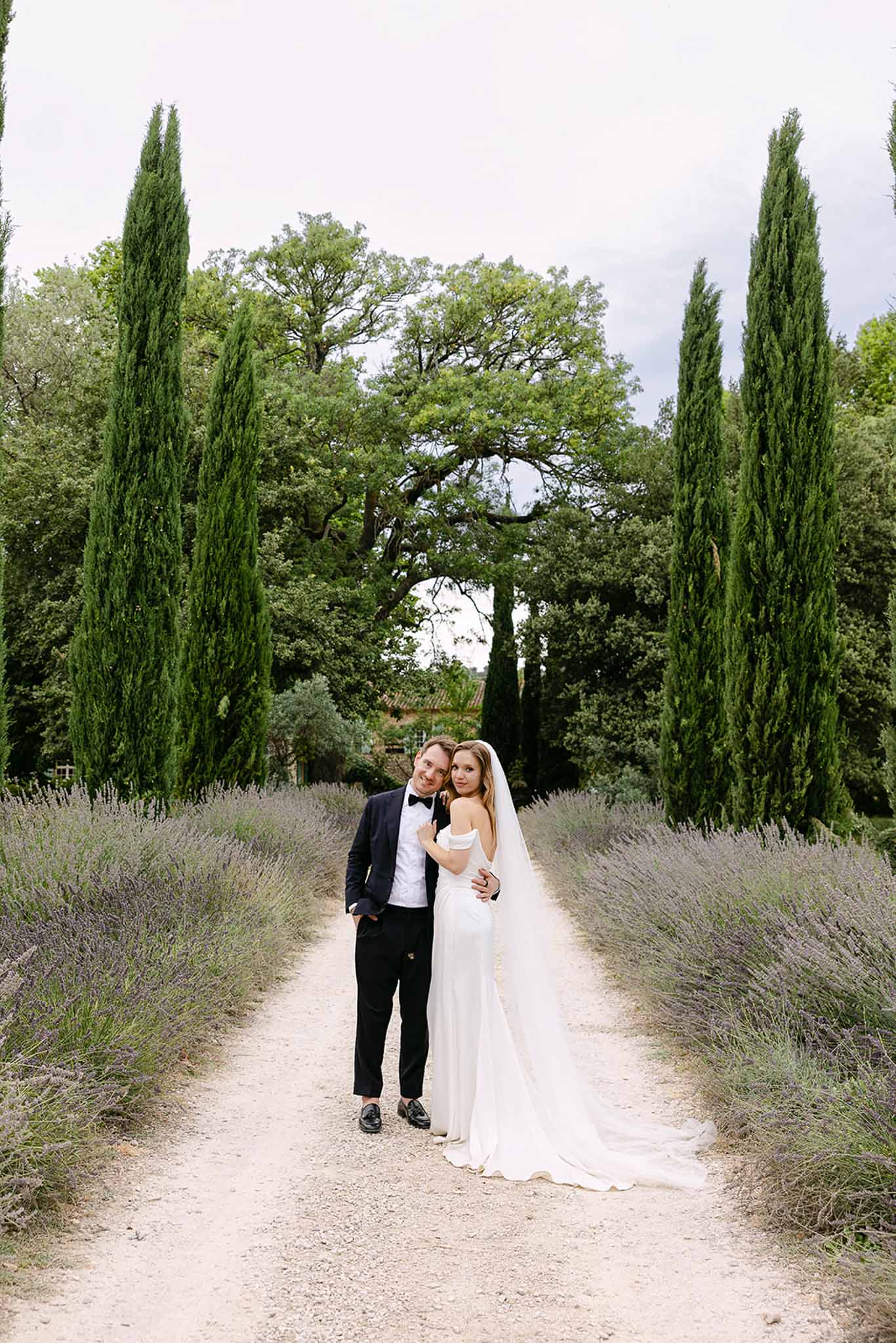 Bride and groom standing on lavender-lined gravel path with cypress trees in Provencal garden