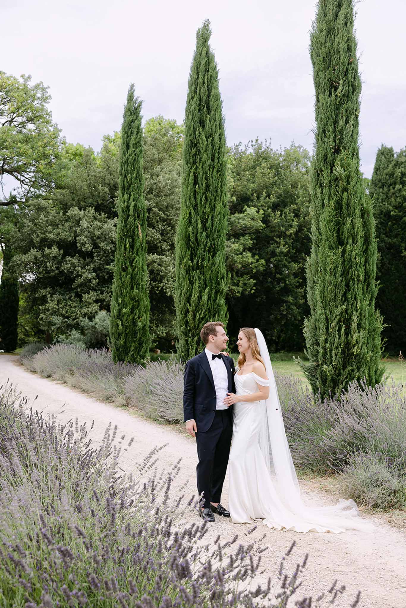 Bride in off-shoulder gown with cathedral veil and groom in navy tuxedo on lavender-lined cypress path
