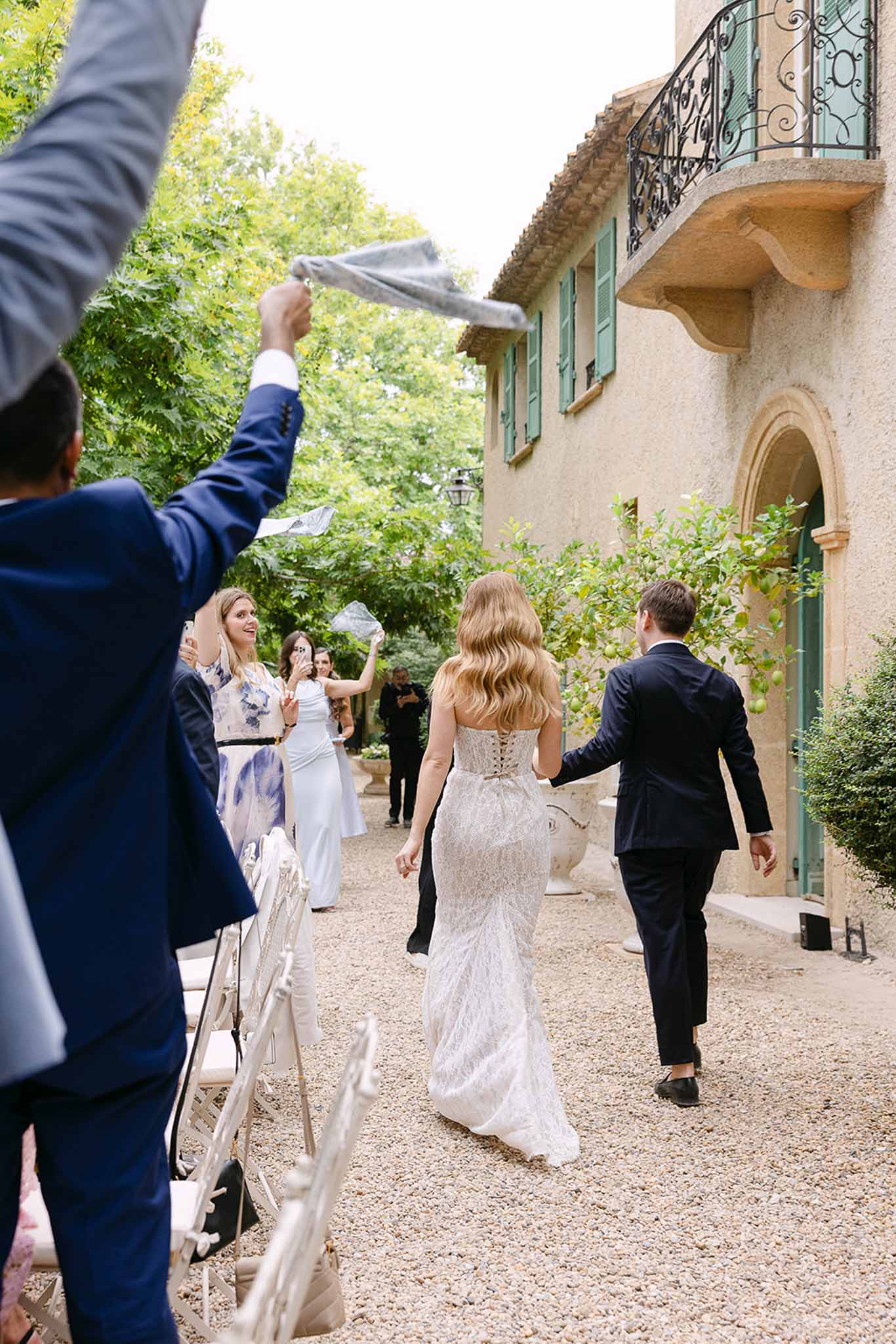Bride and groom walking away as guests wave handkerchiefs at Provencal mas with sage shutters and lemon tree