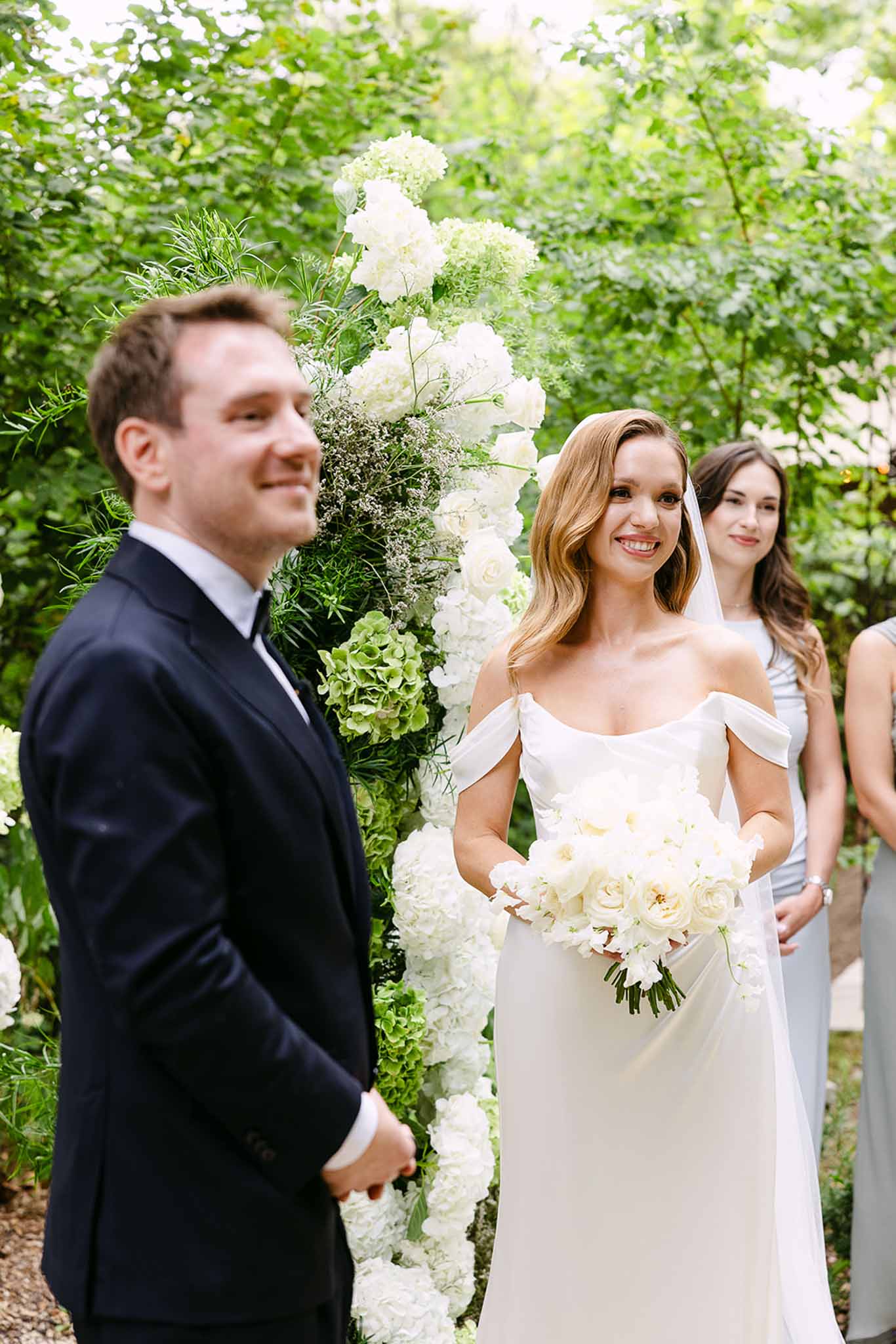 Bride holding white peony bouquet and groom at altar beneath white and green hydrangea arch