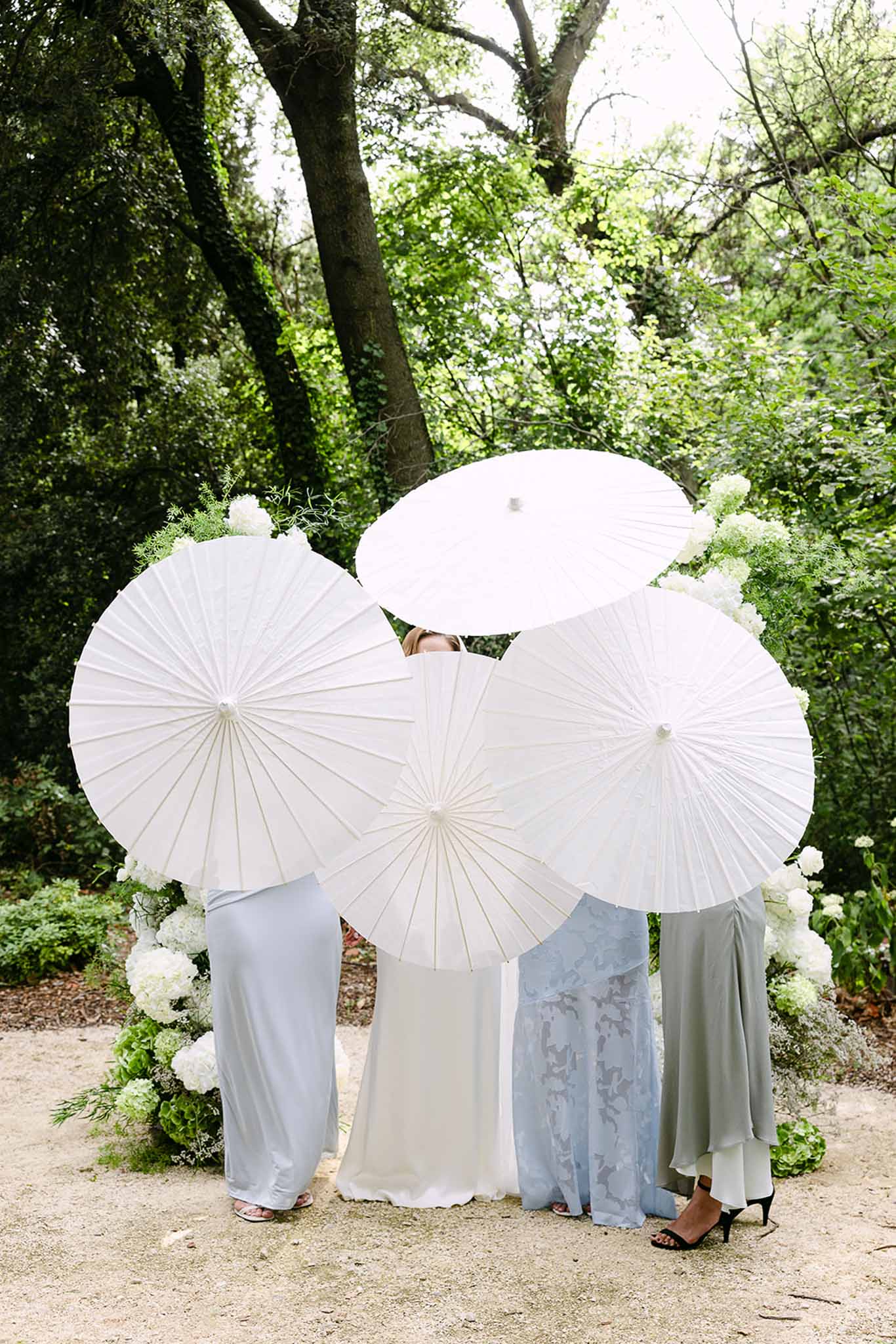 Bride and three bridesmaids in pastel dresses holding white parasols over faces in garden