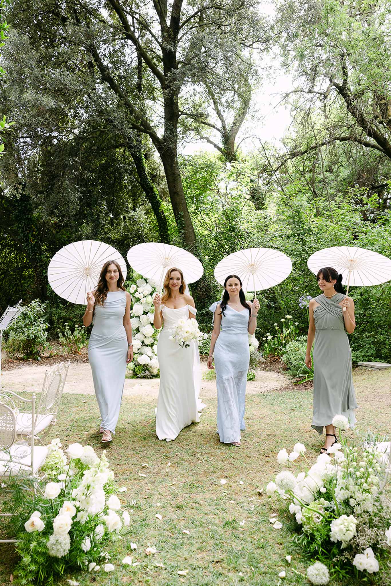 Bride and three bridesmaids walking down outdoor garden aisle holding white parasols with white peony arrangements lining ...