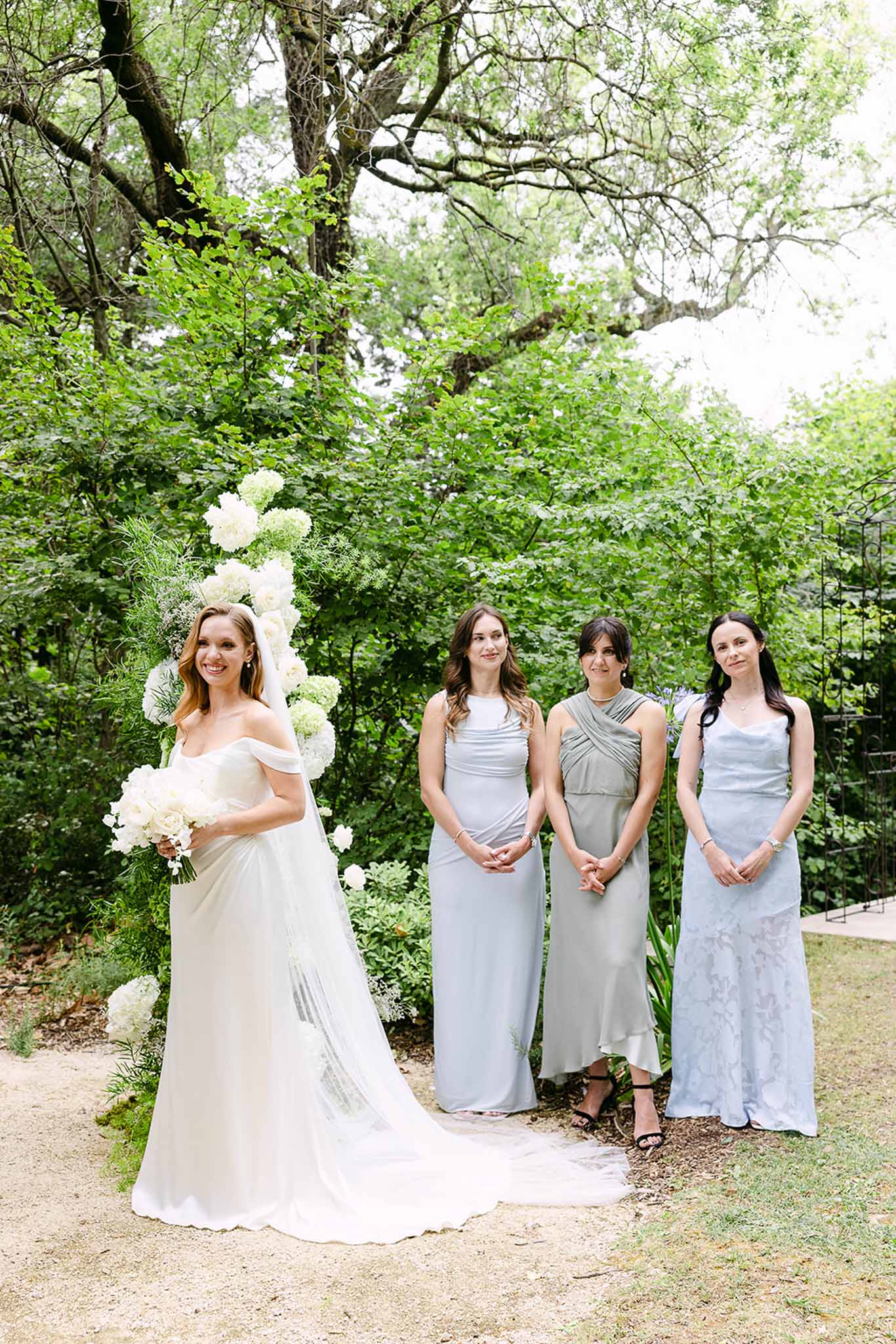 Bride with cathedral veil and three bridesmaids in pale blue and sage beside white hydrangea floral column in garden