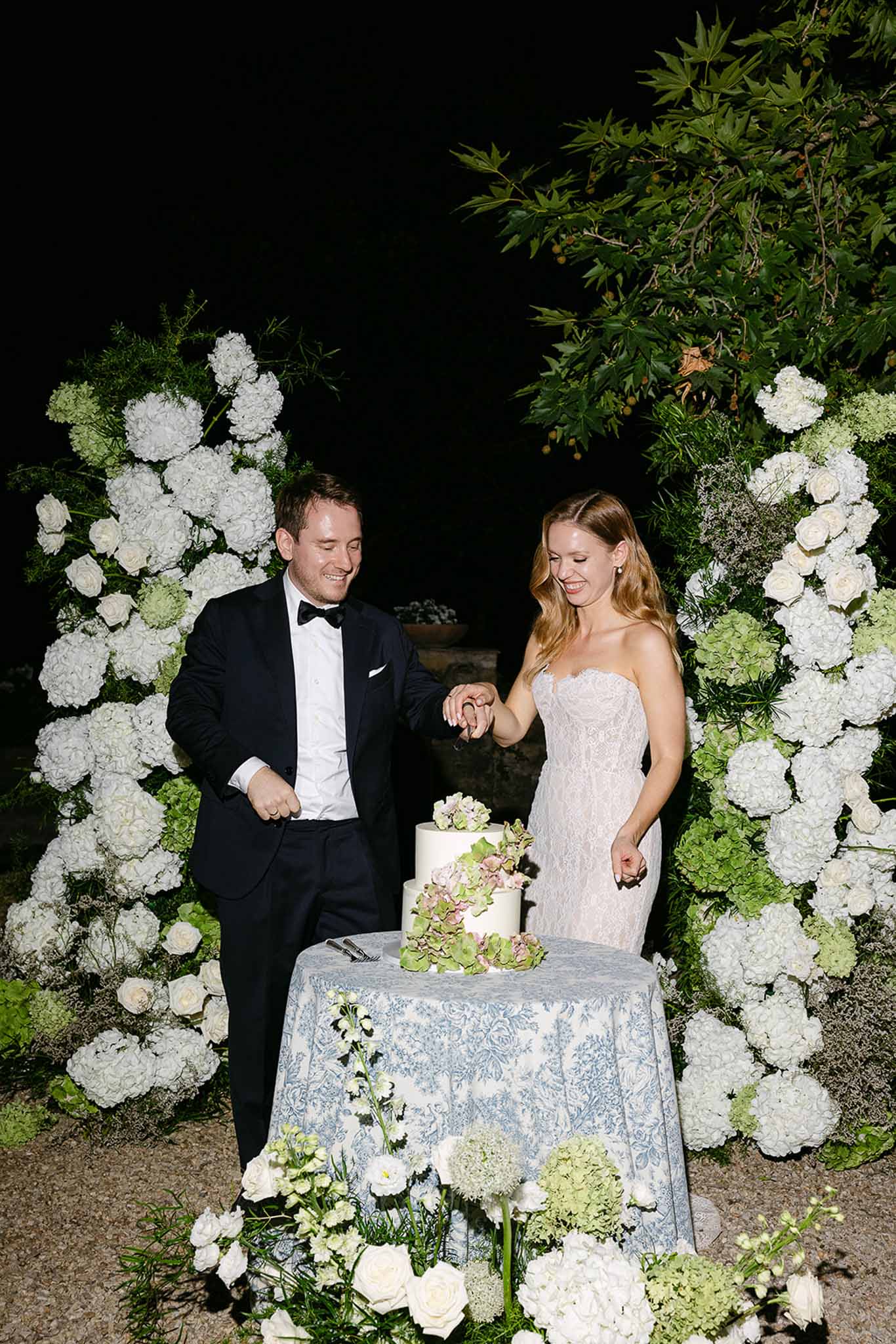 Bride and groom cutting three-tier white wedding cake flanked by hydrangea floral columns at evening reception