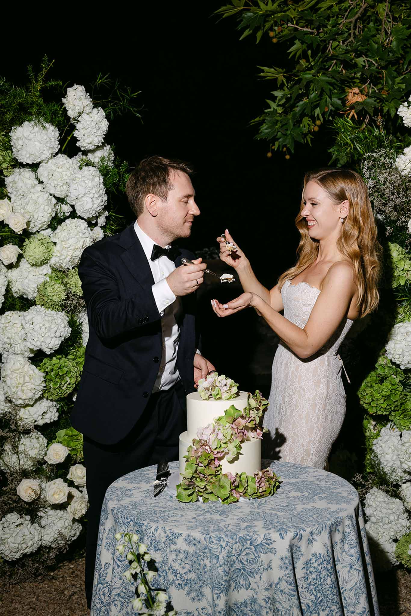 Couple cutting two-tier white cake on toile de Jouy linen with trailing hydrangea and white floral backdrop at night