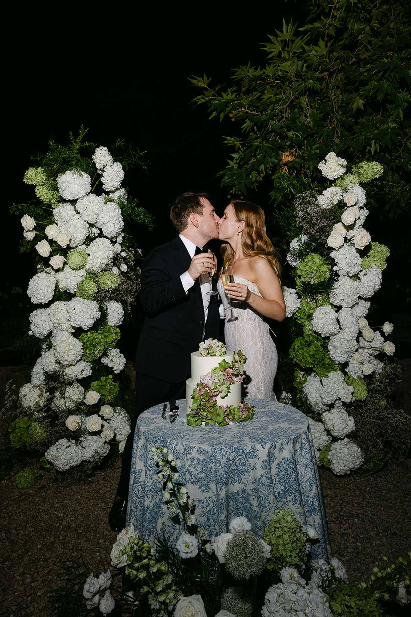 Bride and groom kiss at cake cutting beside three-tier white cake flanked by hydrangea columns at night