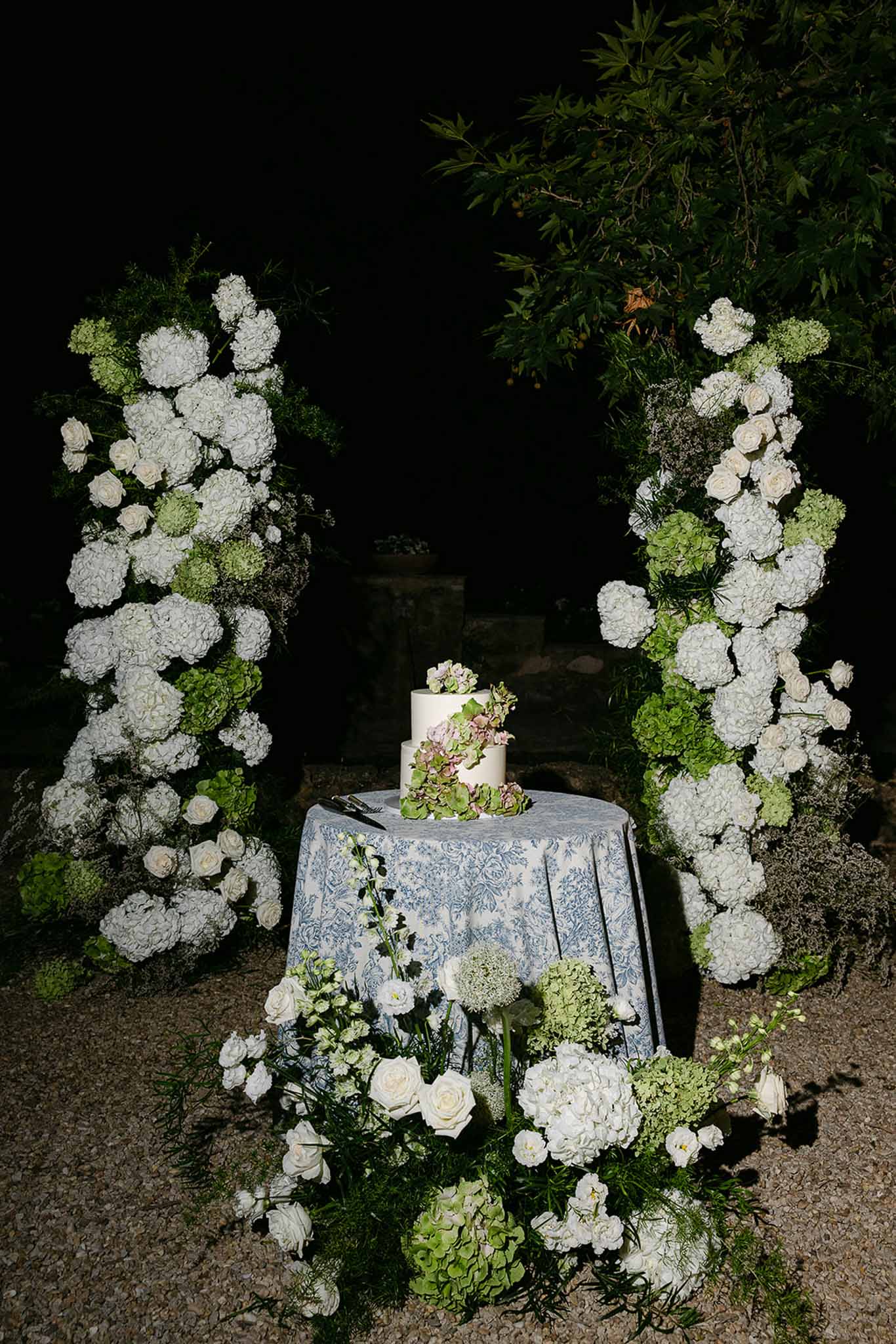 Three-tiered white wedding cake with hydrangeas flanked by tall white and green floral columns on toile-covered table