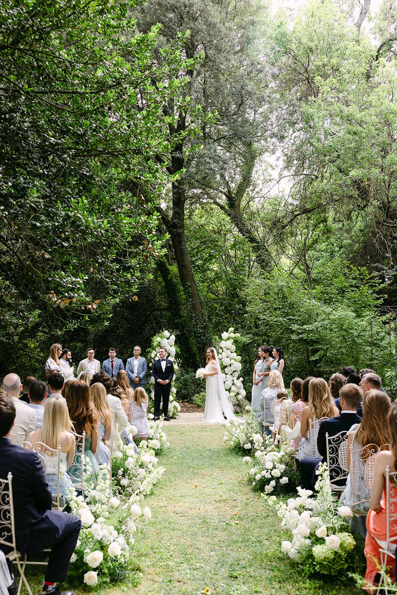 Garden ceremony with all-white rose and hydrangea floral columns sixty guests bridesmaids in sage and groomsmen at altar