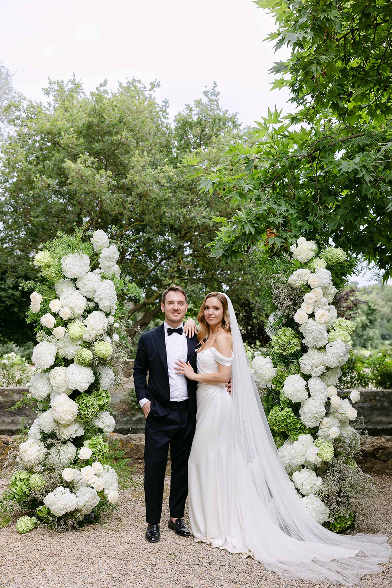 Bride in ivory satin gown with veil and groom in tuxedo between white hydrangea and rose floral columns
