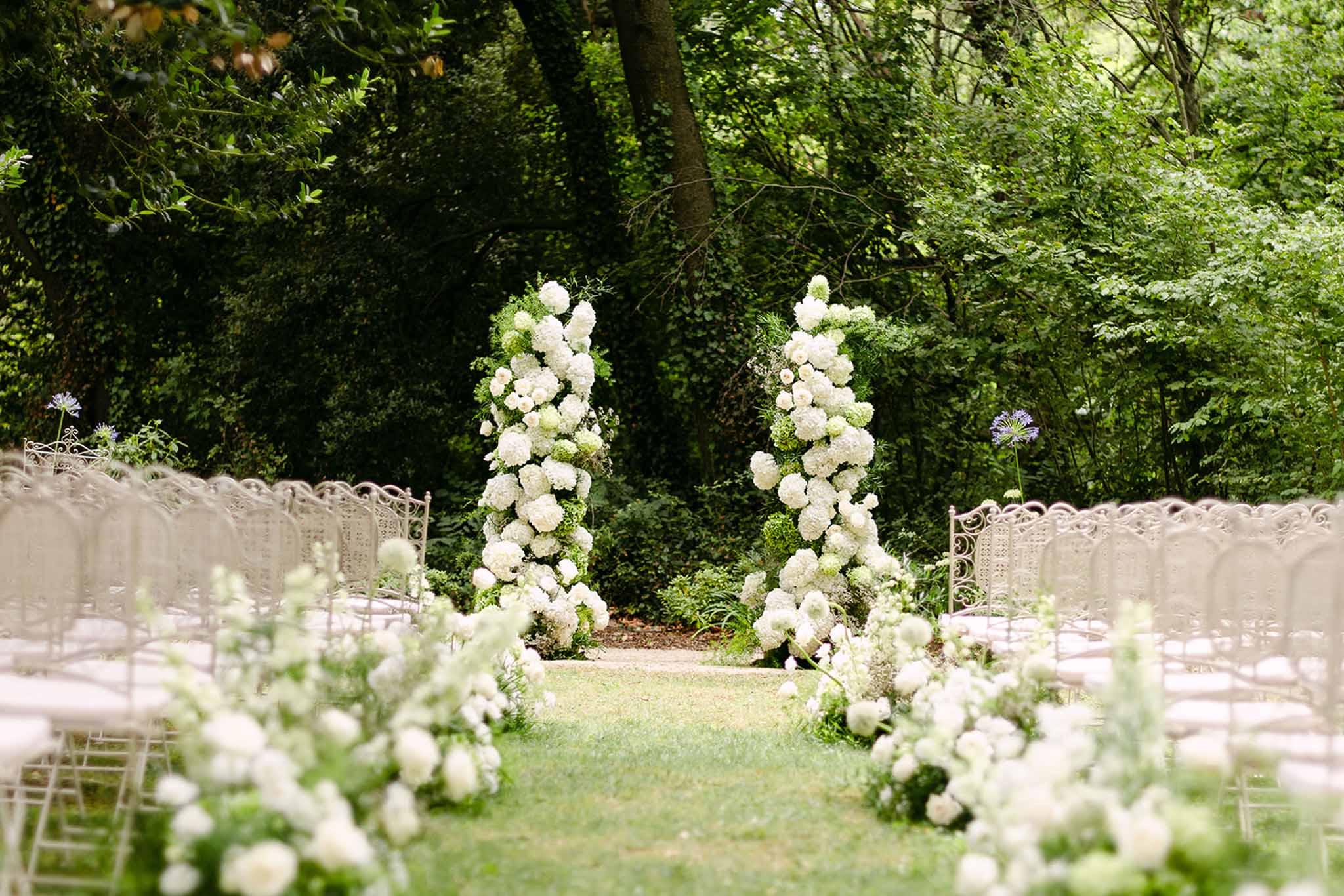 White hydrangea columns framing altar with white rose aisle arrangements and wrought-iron chairs in wooded garden