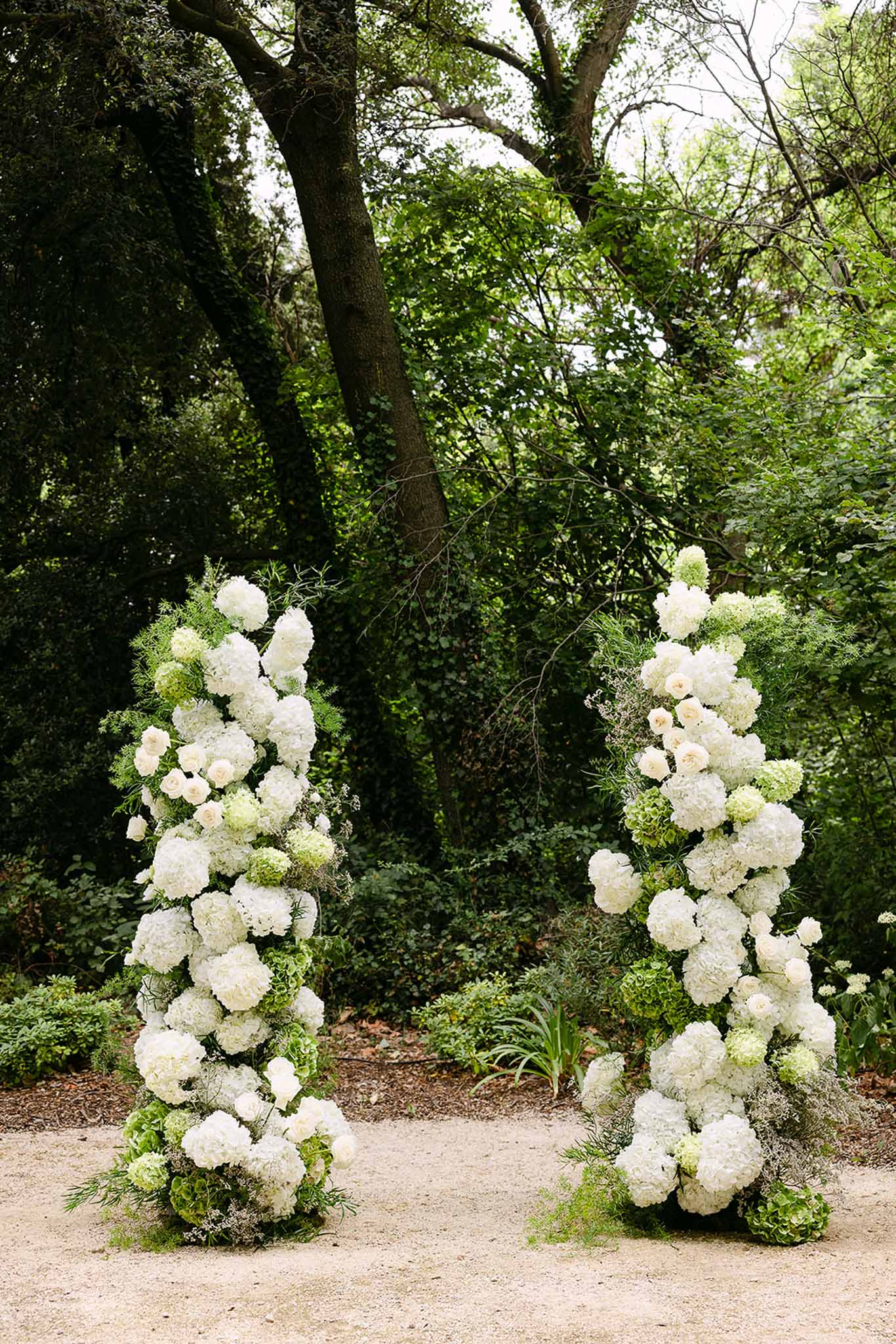 Two freestanding floral column arrangements of white hydrangeas and garden roses on gravel path