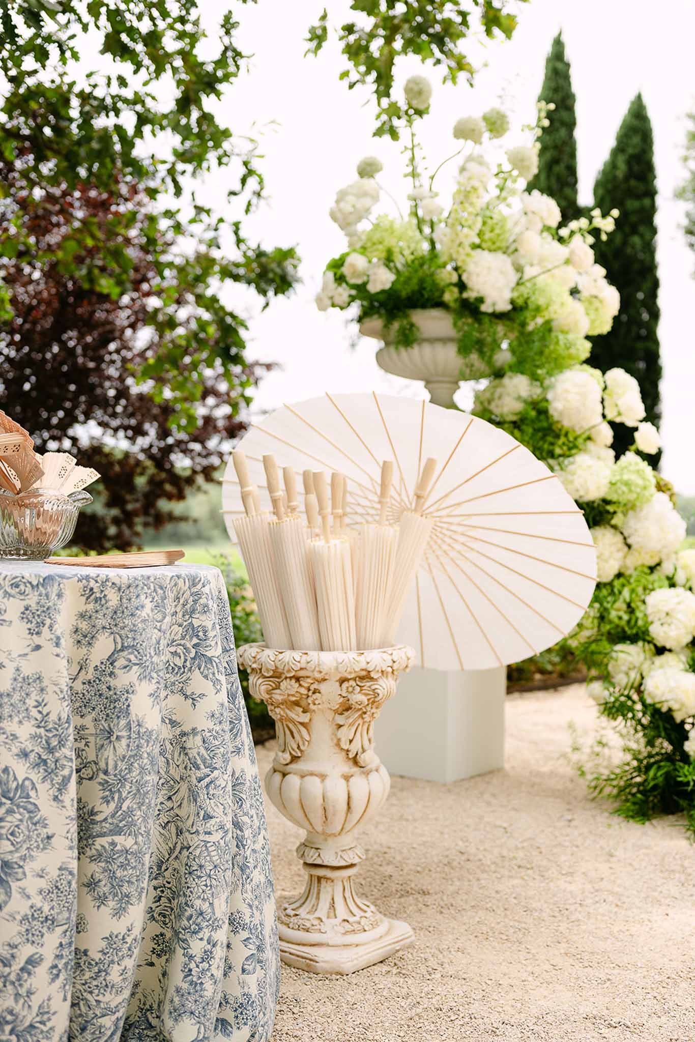 Guest amenities station with paper parasols and toile de Jouy linen beside stone urns of white hydrangeas in a French garden