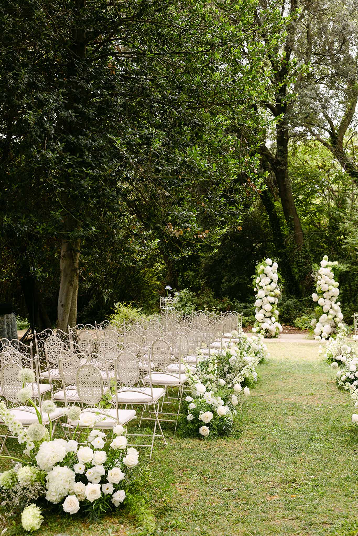 White garden ceremony setup with wrought-iron chairs, rose aisle borders, and tall hydrangea column archway