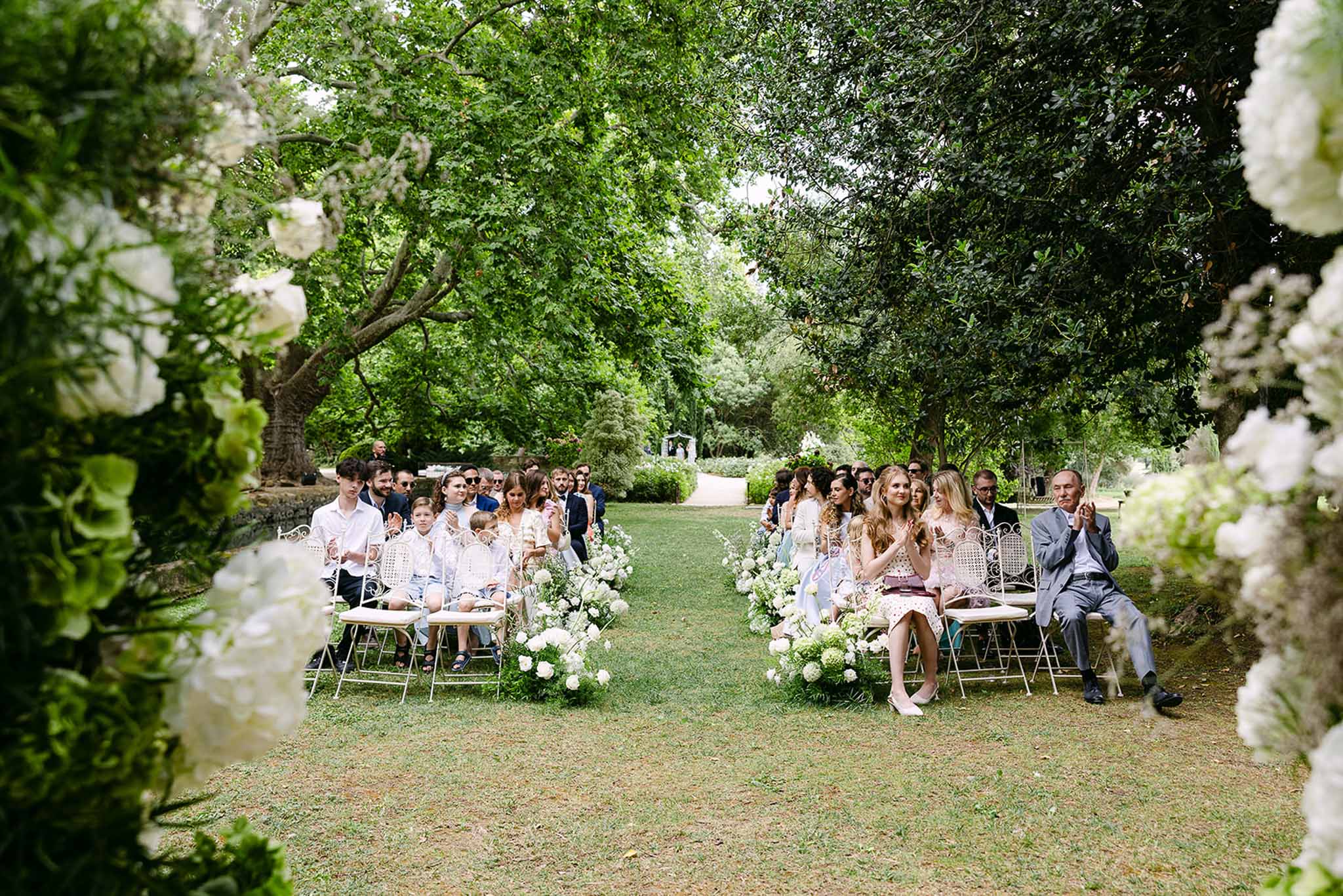 Outdoor ceremony with guests seated on white chairs in tree-lined garden, white hydrangea aisle arrangements