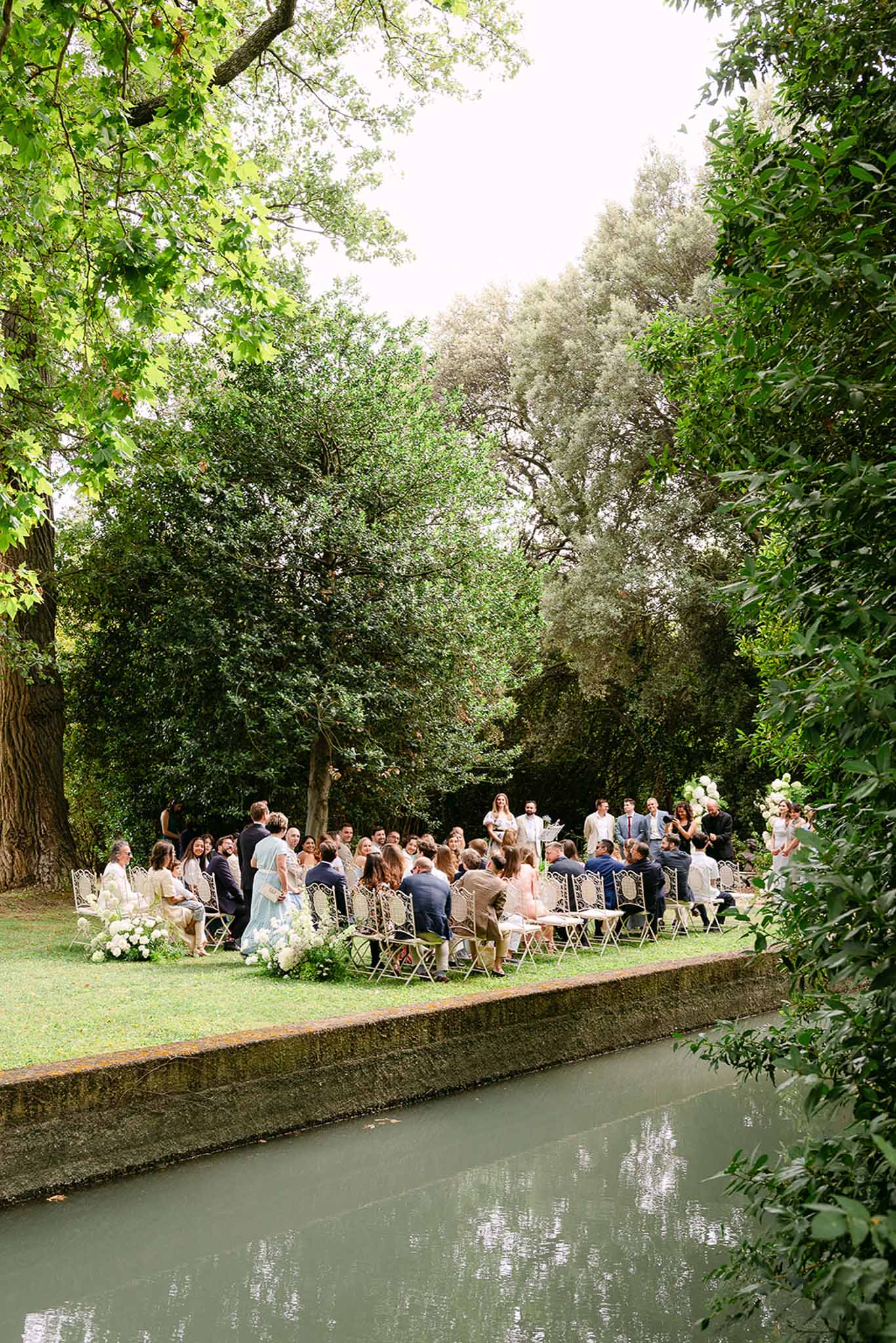 Outdoor wedding ceremony beside a canal with guests on gold chairs, white hydrangea aisle decor and mature tree canopy