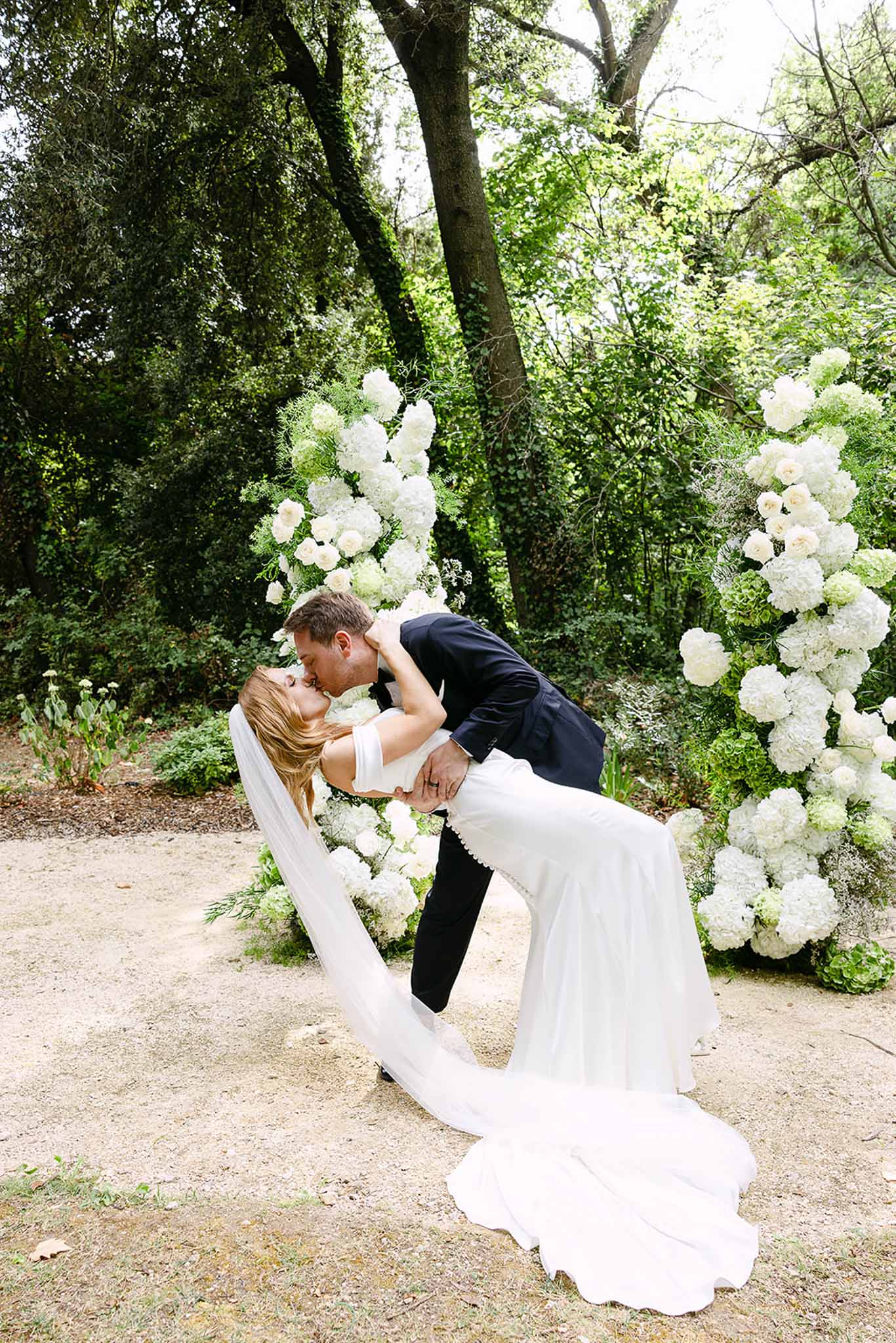 Bride and groom sharing a dip kiss in front of a circular white hydrangea and rose floral arch in a garden